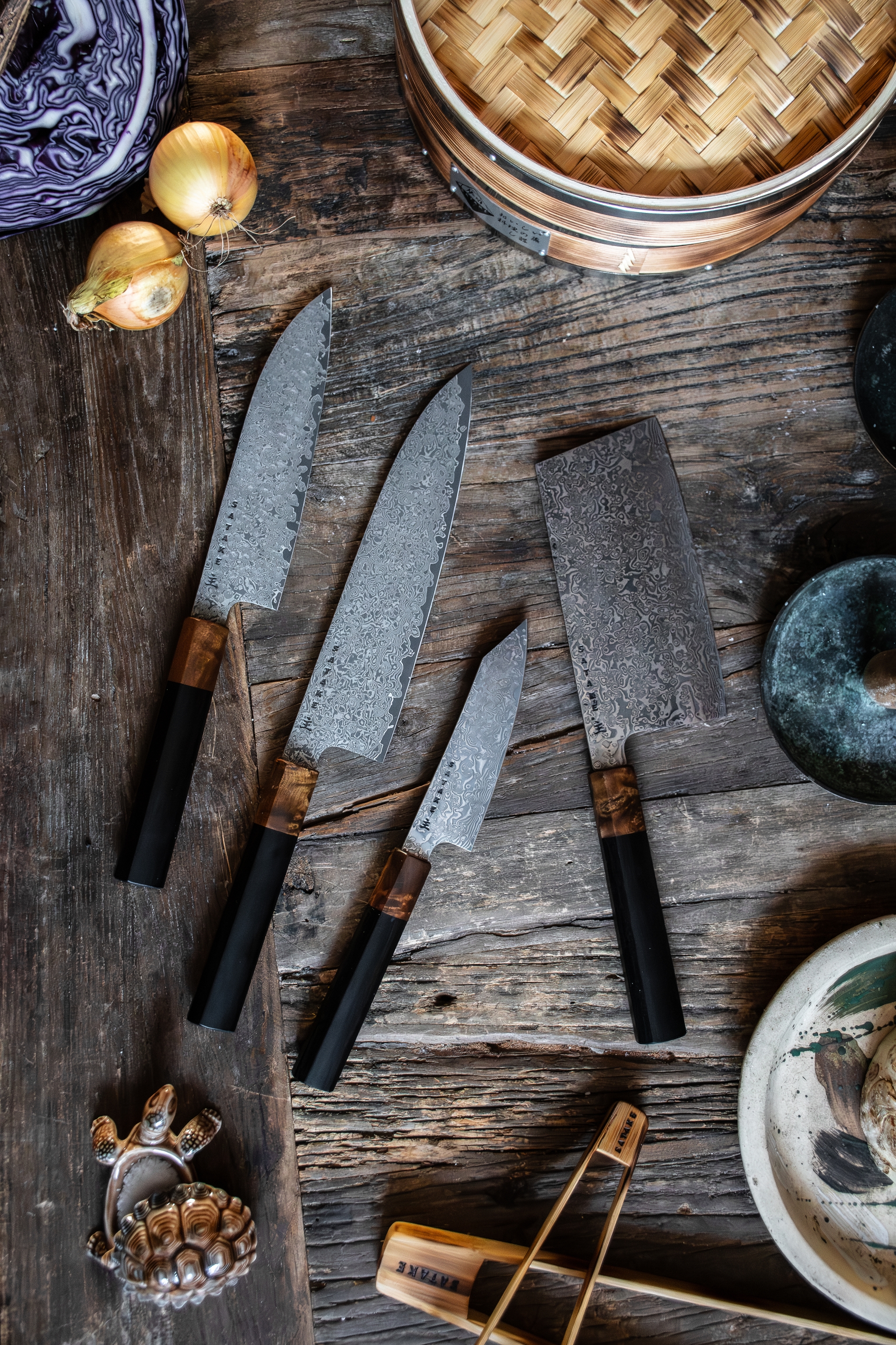 Fyra köksknivar i damaskusstål, en ångkokare i bambu, lök och kål på ett rustikt träbord.