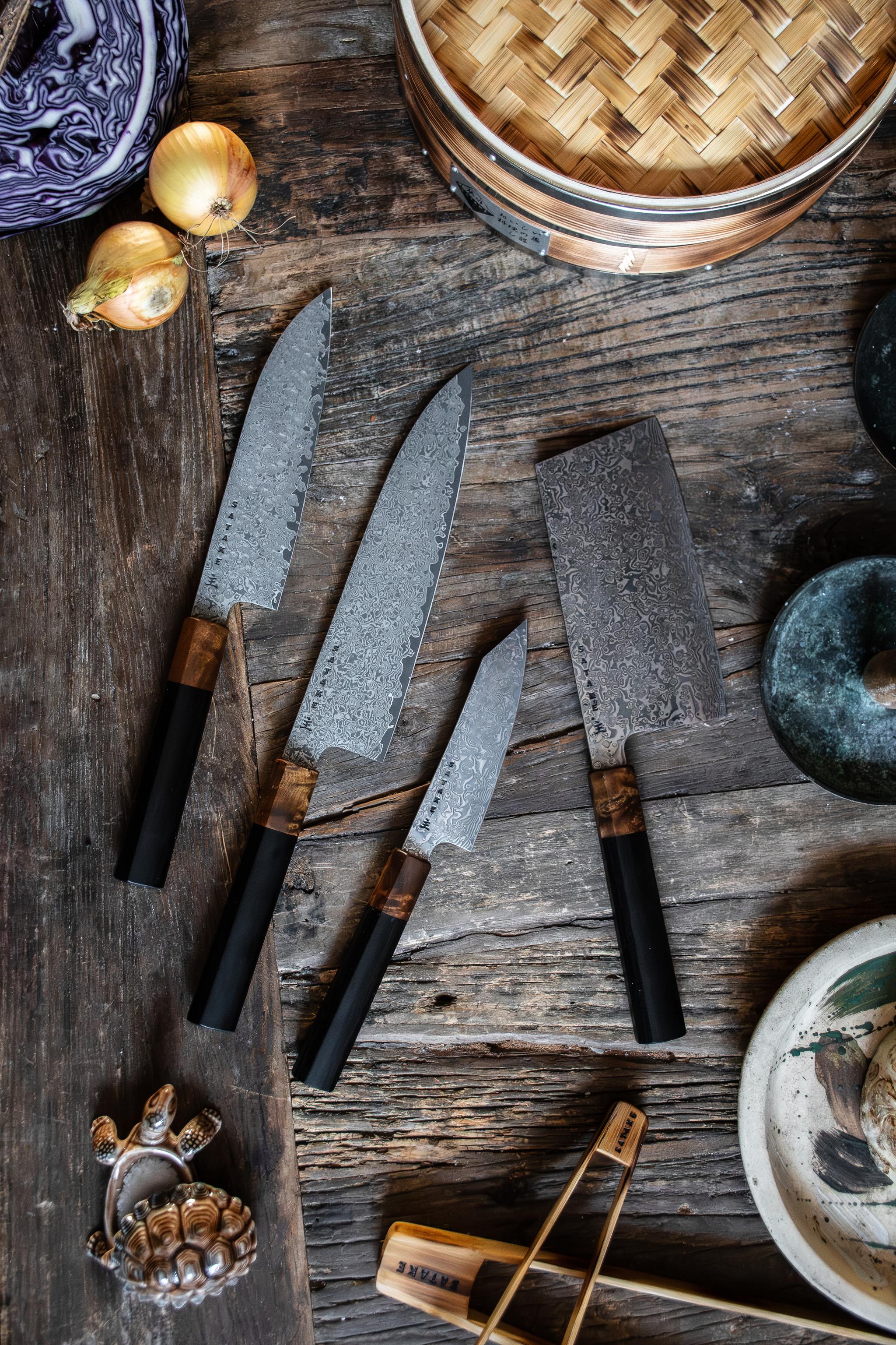 Fyra köksknivar i damaskusstål, en ångkokare i bambu, lök och kål på ett rustikt träbord.