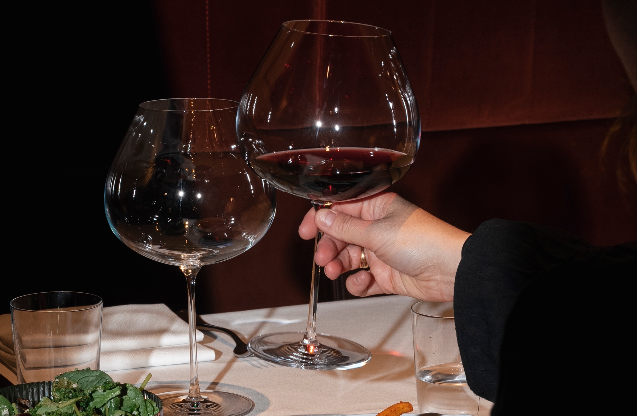 En person håller ett glas rött vin vid ett restaurangbord dukat med pommes frites, kött och sallad.