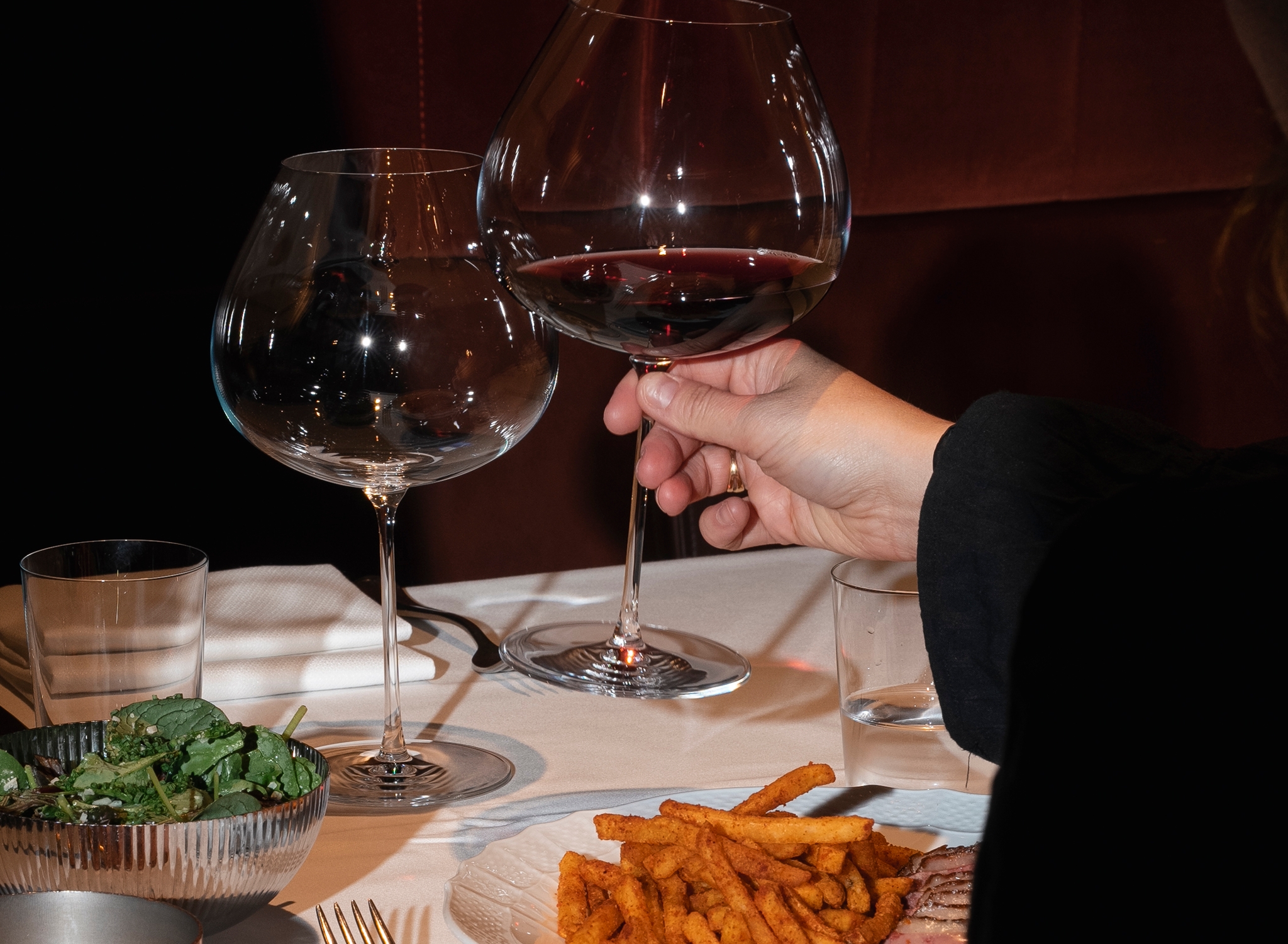 En person håller ett glas rött vin vid ett restaurangbord med pommes frites, kött och sallad.