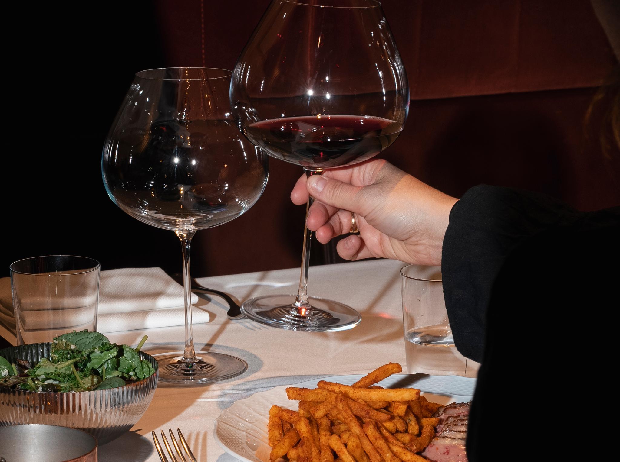 Person håller ett glas rött vin vid ett dukat restaurangbord med pommes frites, kött och sallad.