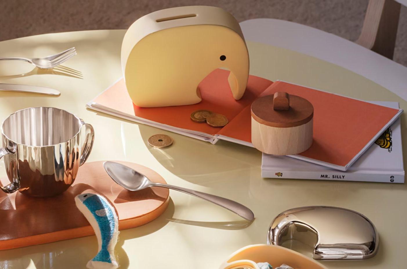 En elefantformad sparbössa, en silverfärgad barnmugg, en leksaksfisk i trä och böcker på ett ljusgult bord
