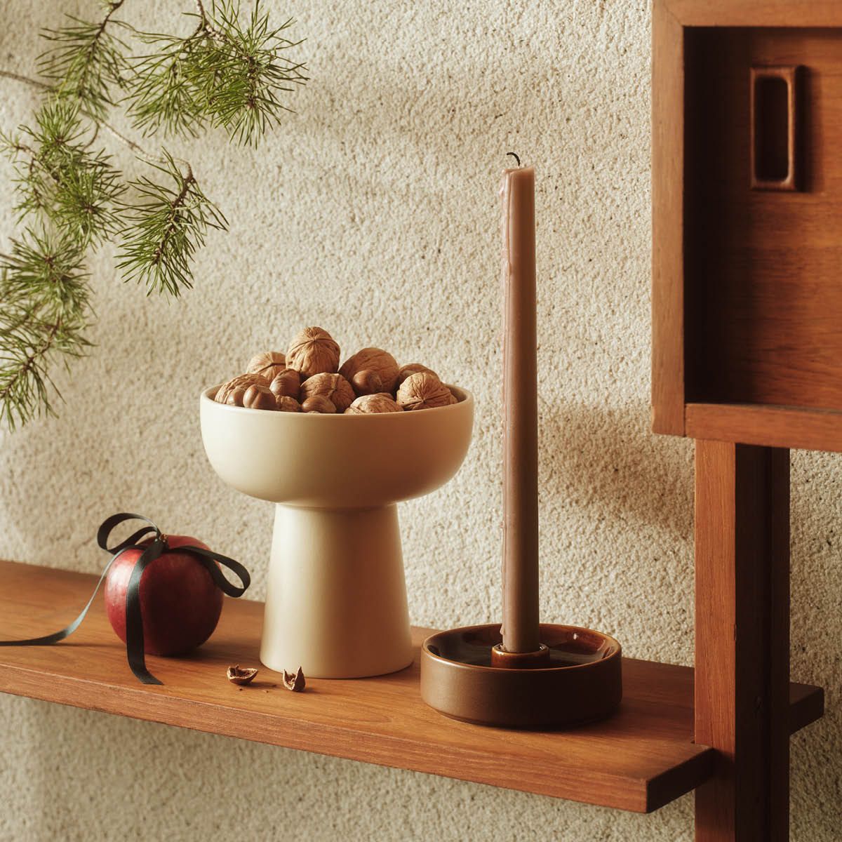 Kermanvärinen jalustakulho täytettynä saksanpähkinöillä, punainen omena mustalla nauhalla ja ruskea kynttilä puisella hyllyllä, männynoksa näkyvissä.
