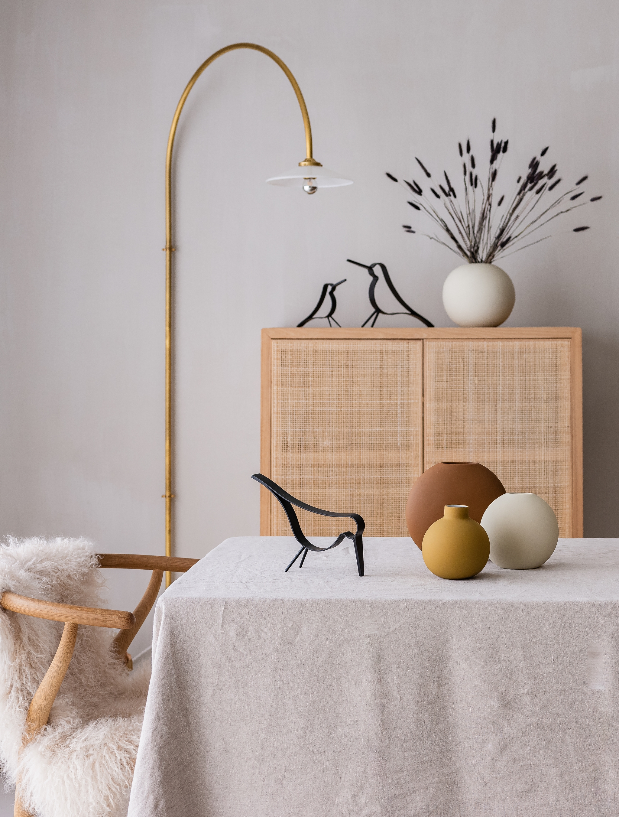 Skandinaviskt inreddt vardagsrum med ljus trästol, linnetäckt bord, runda keramikvaser och minimalistiska fågeldetaljer framför ett skåp i rotting.