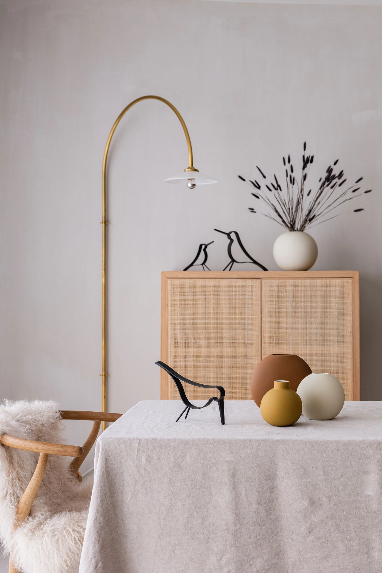 Skandinaviskt inreddt vardagsrum med ljus trästol, linnetäckt bord, runda keramikvaser och minimalistiska fågeldetaljer framför ett skåp i rotting.