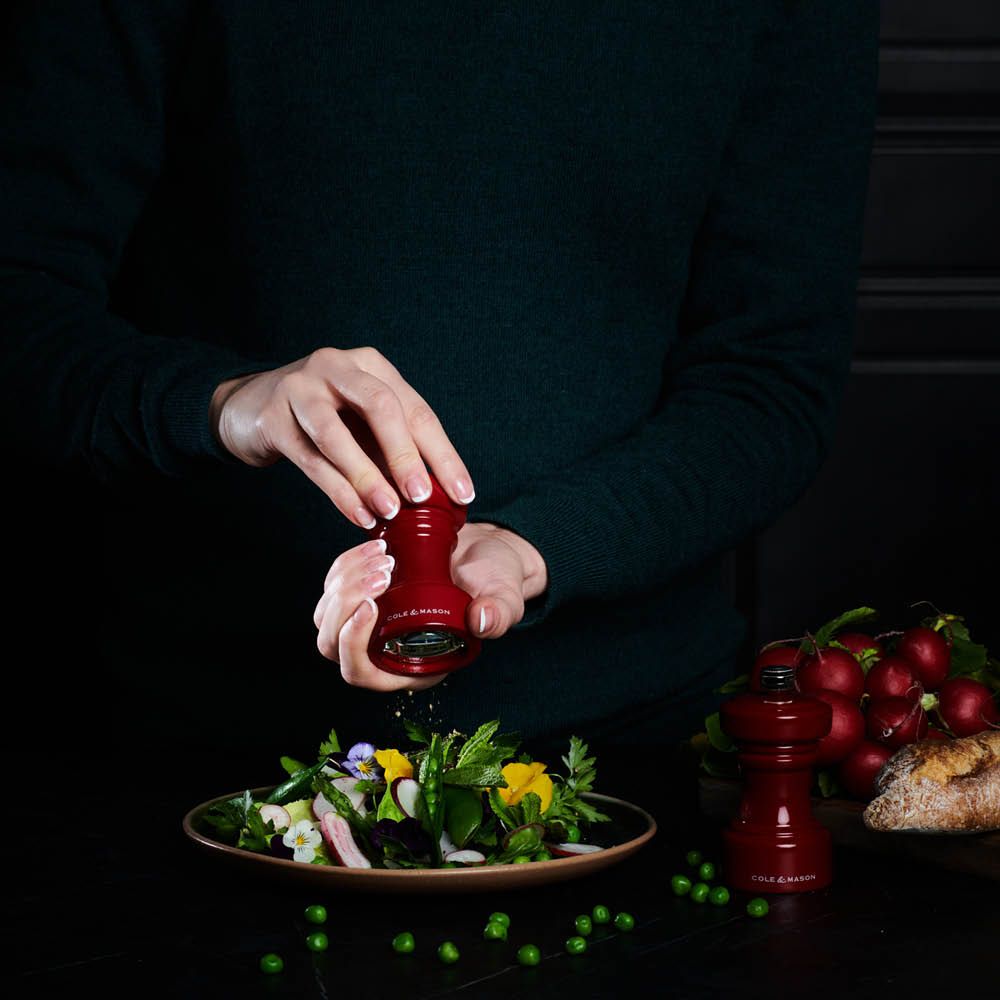 Henkilö maustaa värikästä salaattia punaisella Cole & Mason -maustemyllyllä tummalla taustalla.