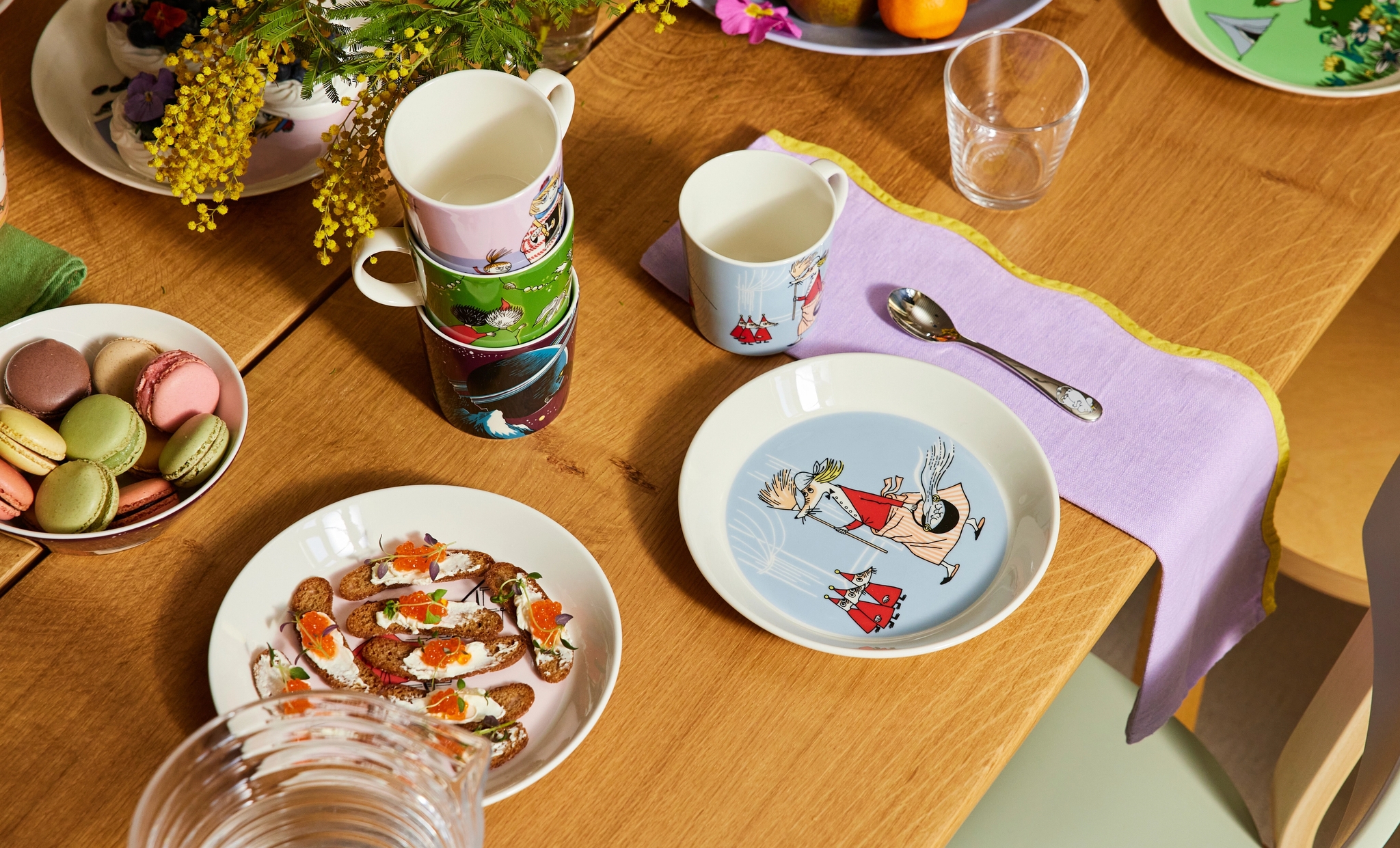 Ett träbord dukat med tallrikar och muggar med Mumintema, färgglada macarons, aptitretare, en fruktskål och gula mimosablommor.