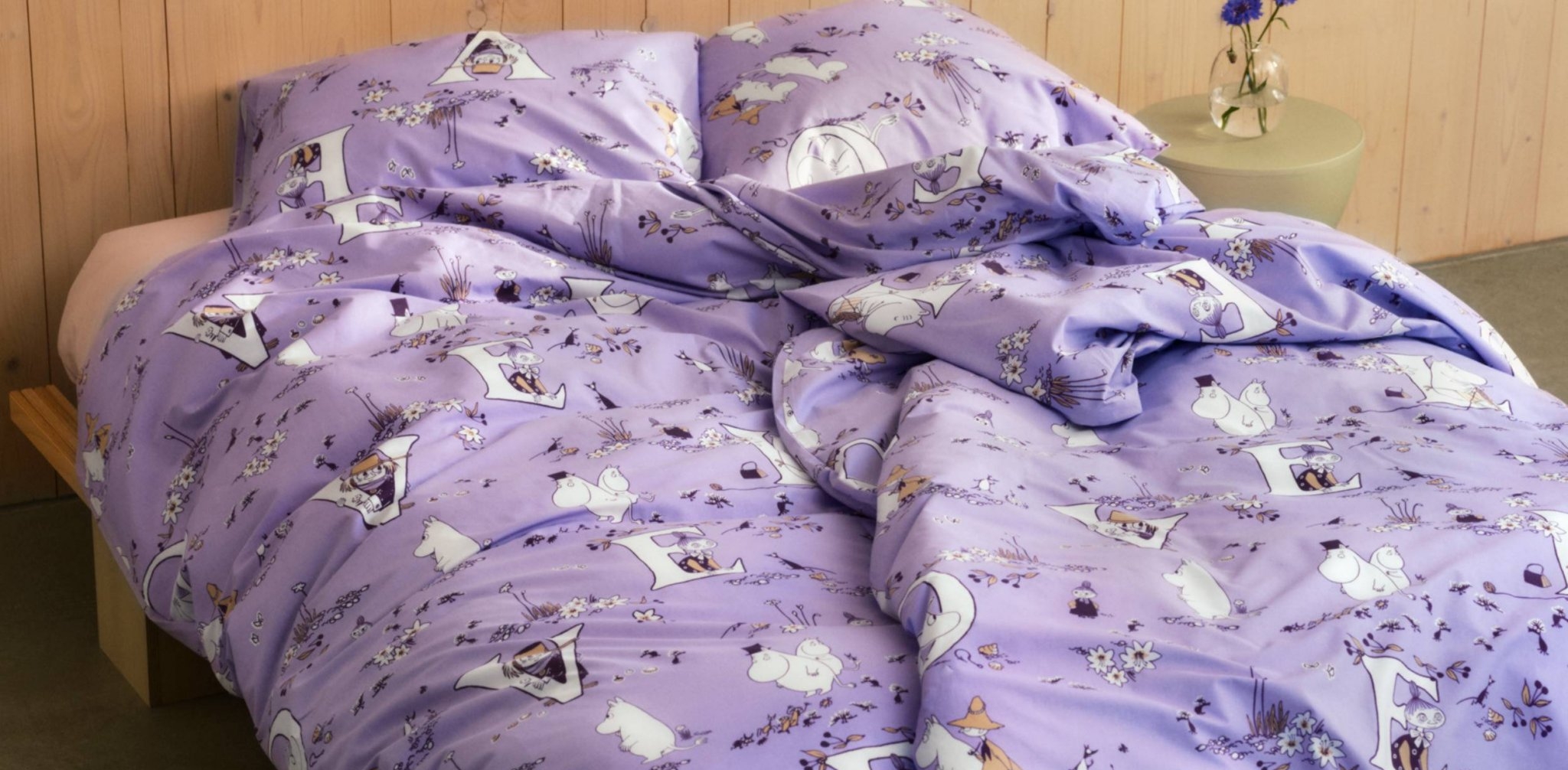 Ljuslila sängkläder med Mumin-tema på en sängram i trä.
