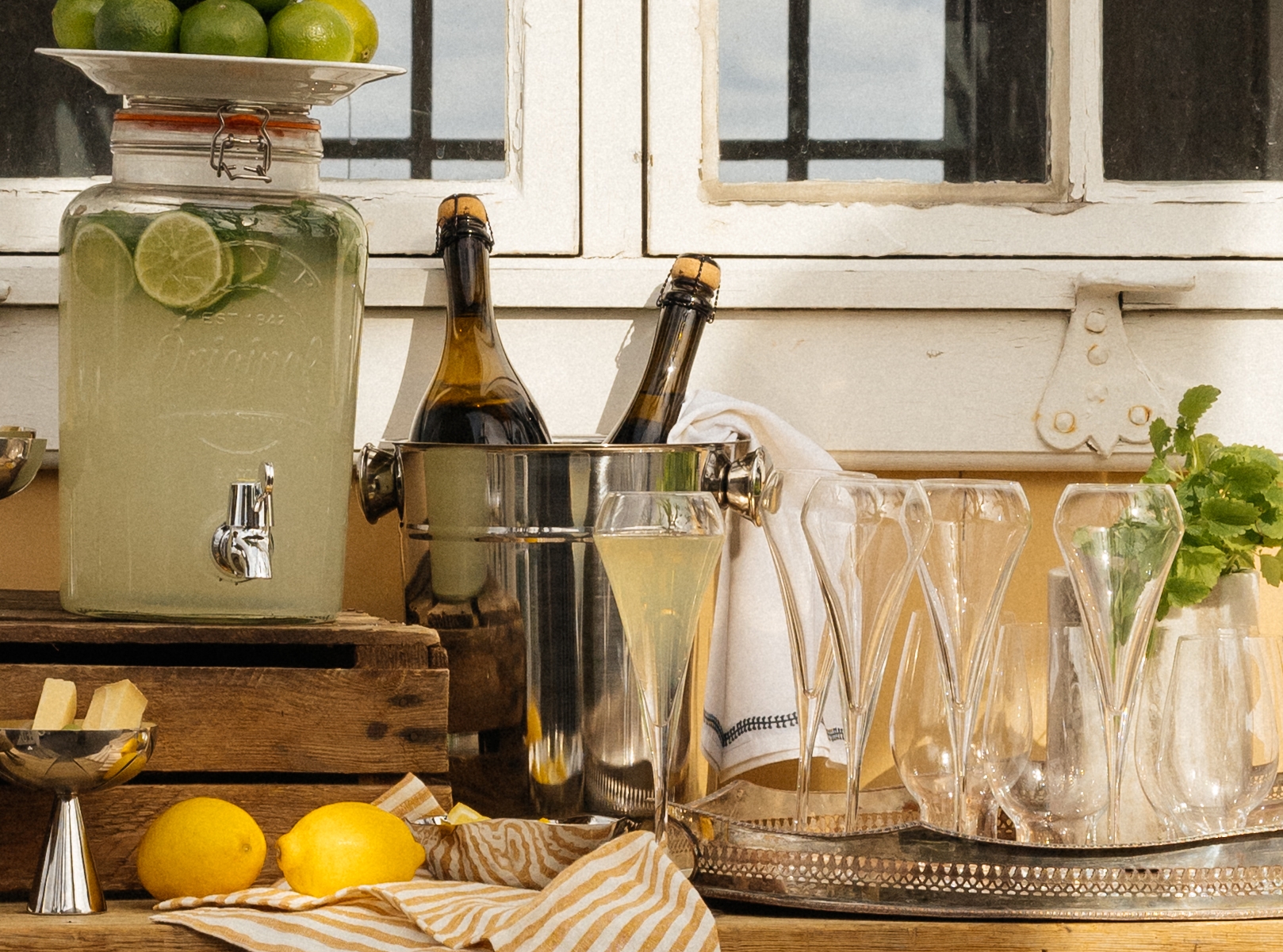 Somrig utomhusdrinkstation med lemonad, kylda flaskor och blommor framför ett gult hus med stora fönster.