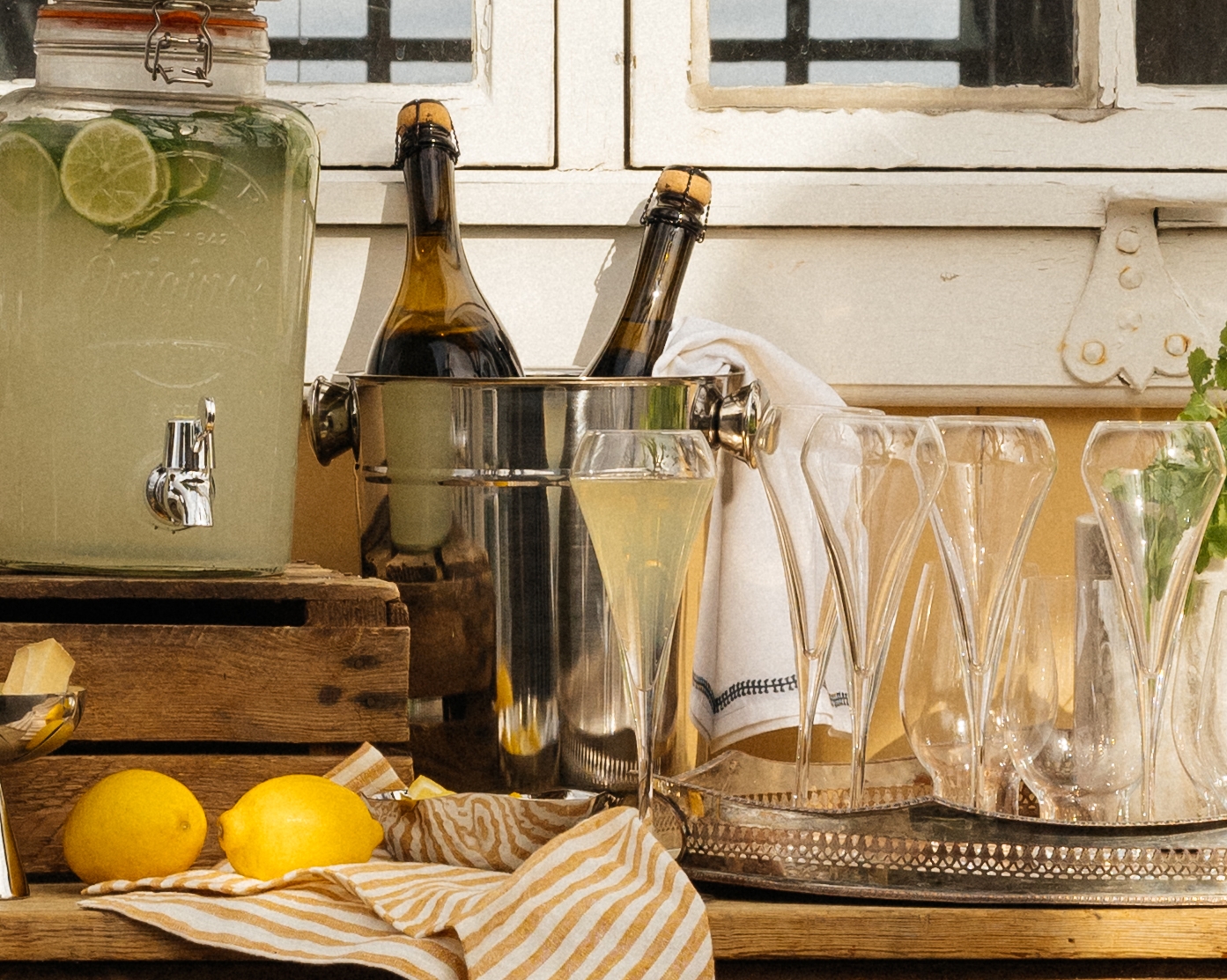 Sommerlig uteservering med sitronlimonade, iskalde drikker og blomster foran et gult hus med store vinduer.