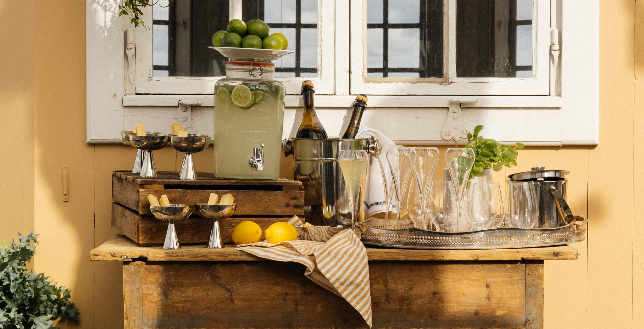 Somrig utomhusbar med lemonad, kylda drycker och gröna växter framför ett gult hus med vita spröjsade fönster.