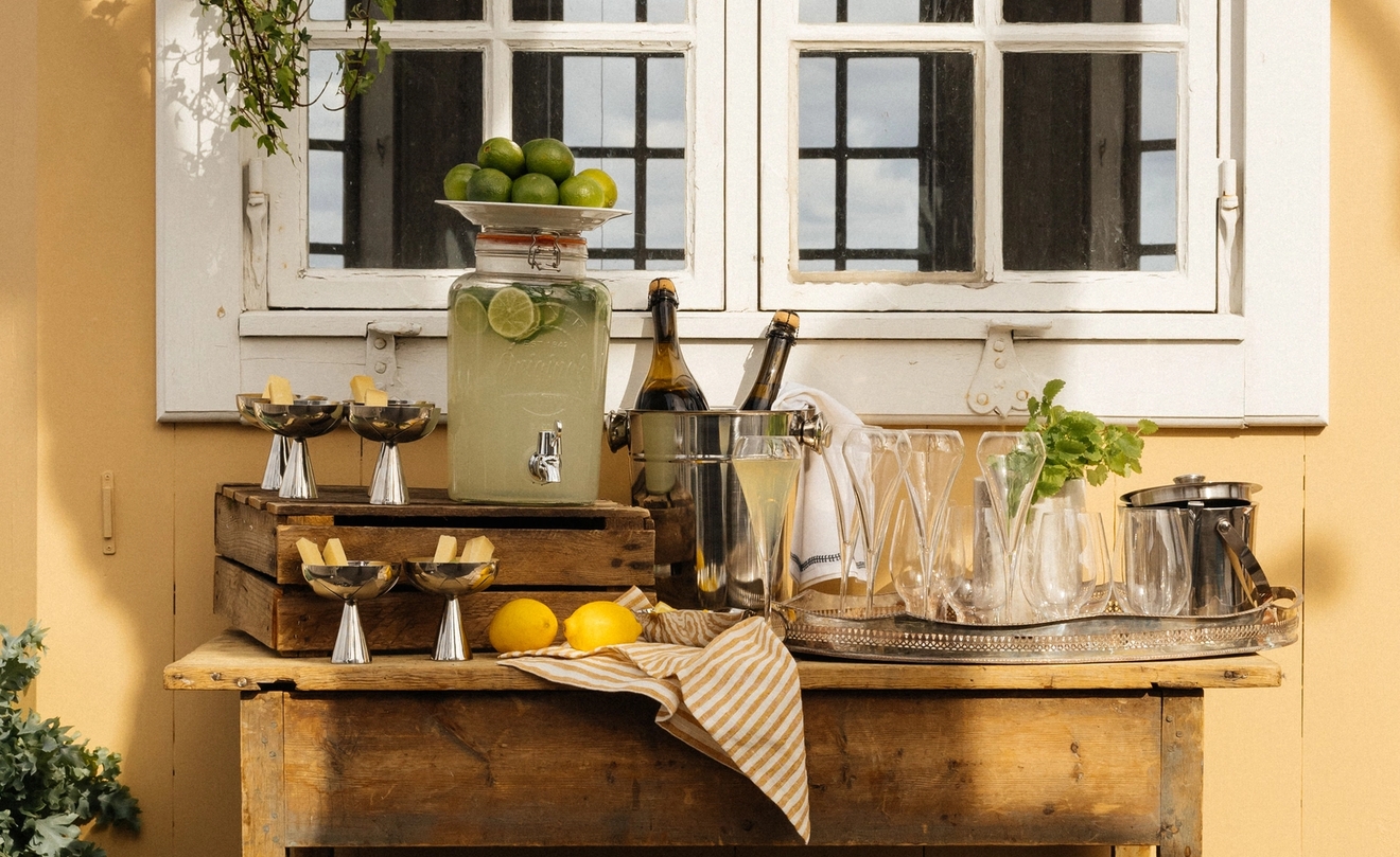 Somrig utomhusdrinkstation med lemonad, flaskor på is och blommor framför ett gult hus med vita fönster.