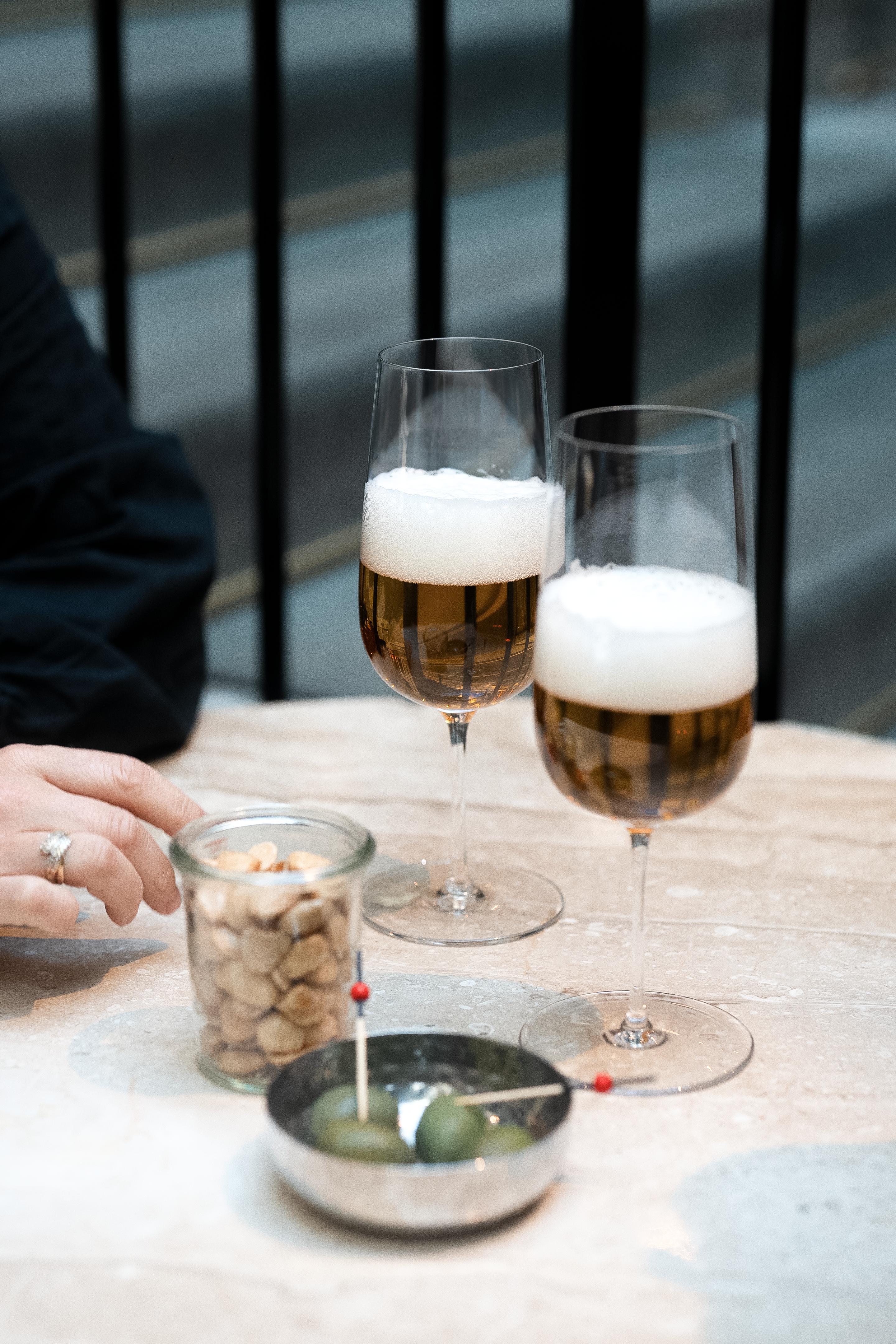 Två glas öl, en burk nötter och en skål oliver på ett ljust bord.