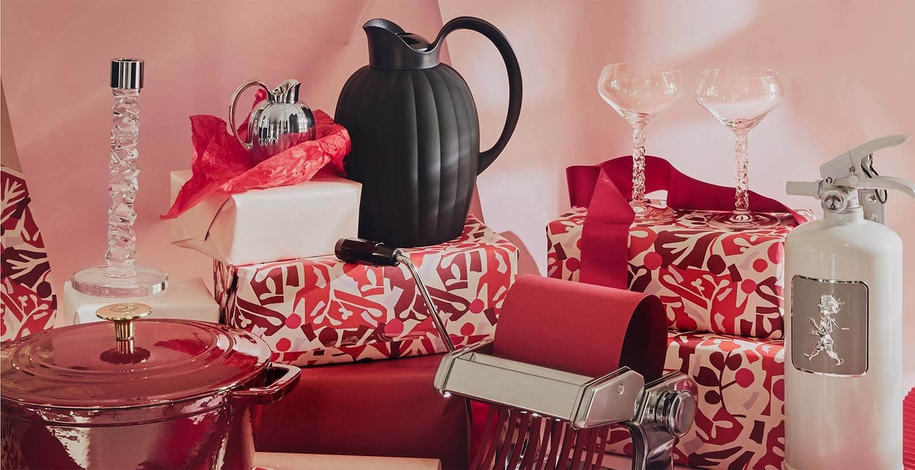Dekorative gjenstander og gaver, inkludert en krystalllysestake, mugger, champagnekupéer og et hvitt brannslukningsapparat, mot en rosa bakgrunn.