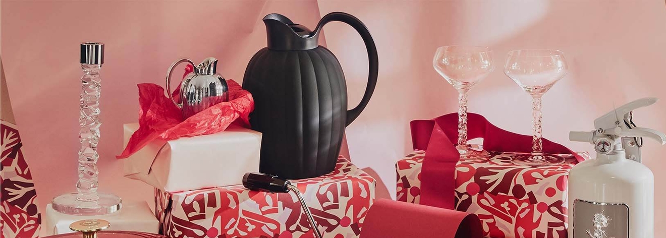Dekorative gjenstander og gaver, inkludert en krystalllysestake, mugger, champagnekupéer og et hvitt brannslukningsapparat, mot en rosa bakgrunn.
