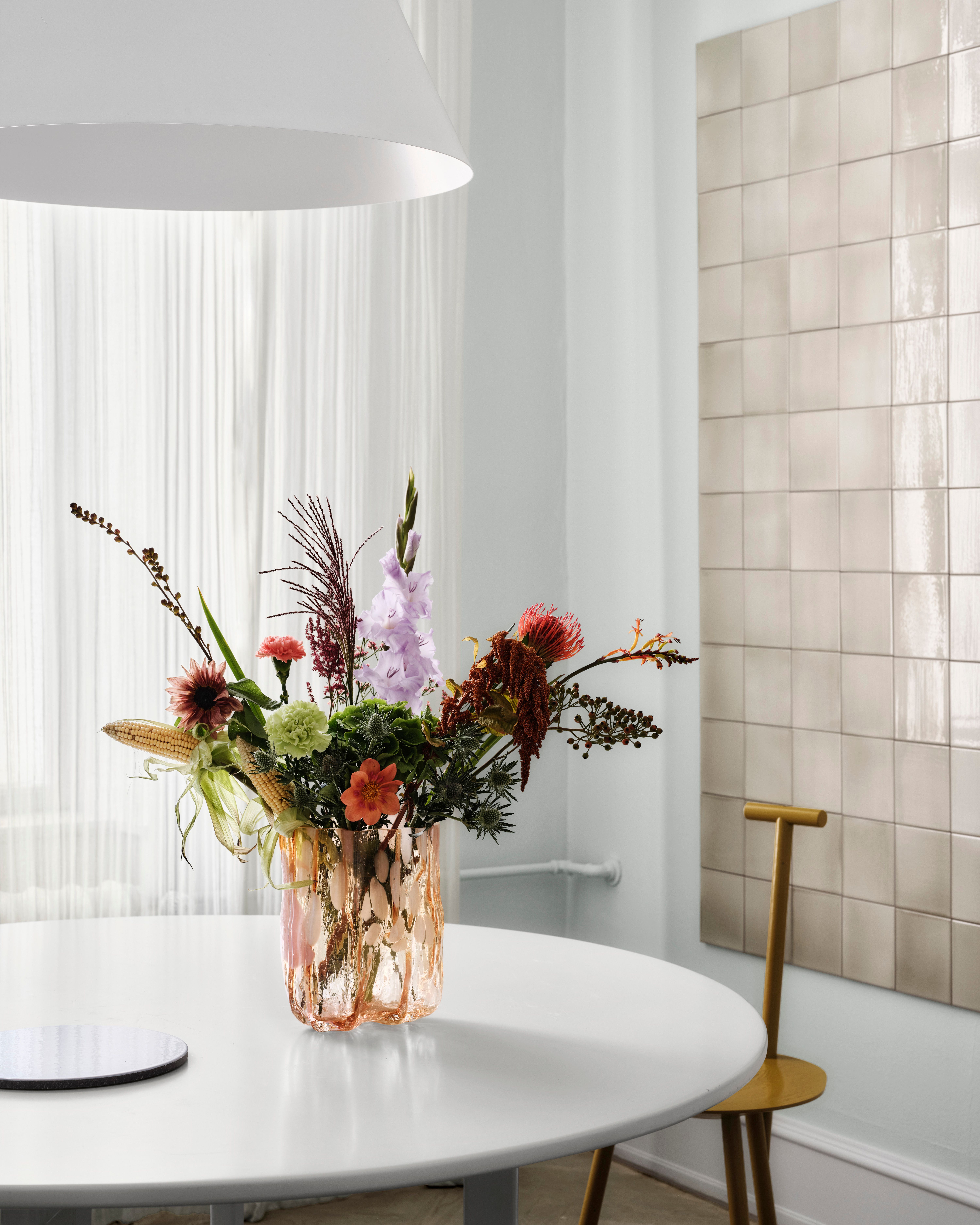 Ett eklektiskt blomsterarrangemang med olika blommor, bladverk och små majskolvar i en texturerad persikrosa vas på ett vitt bord.