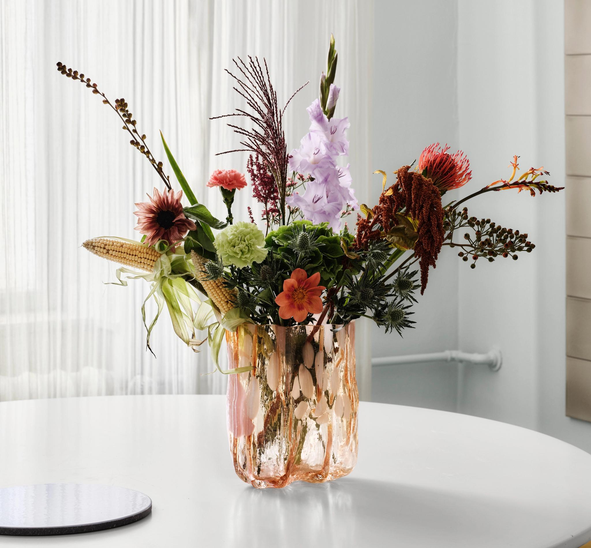 Ett eklektiskt blomsterarrangemang med olika blommor, bladverk och små majskolvar i en texturerad persikrosa vas på ett vitt bord.