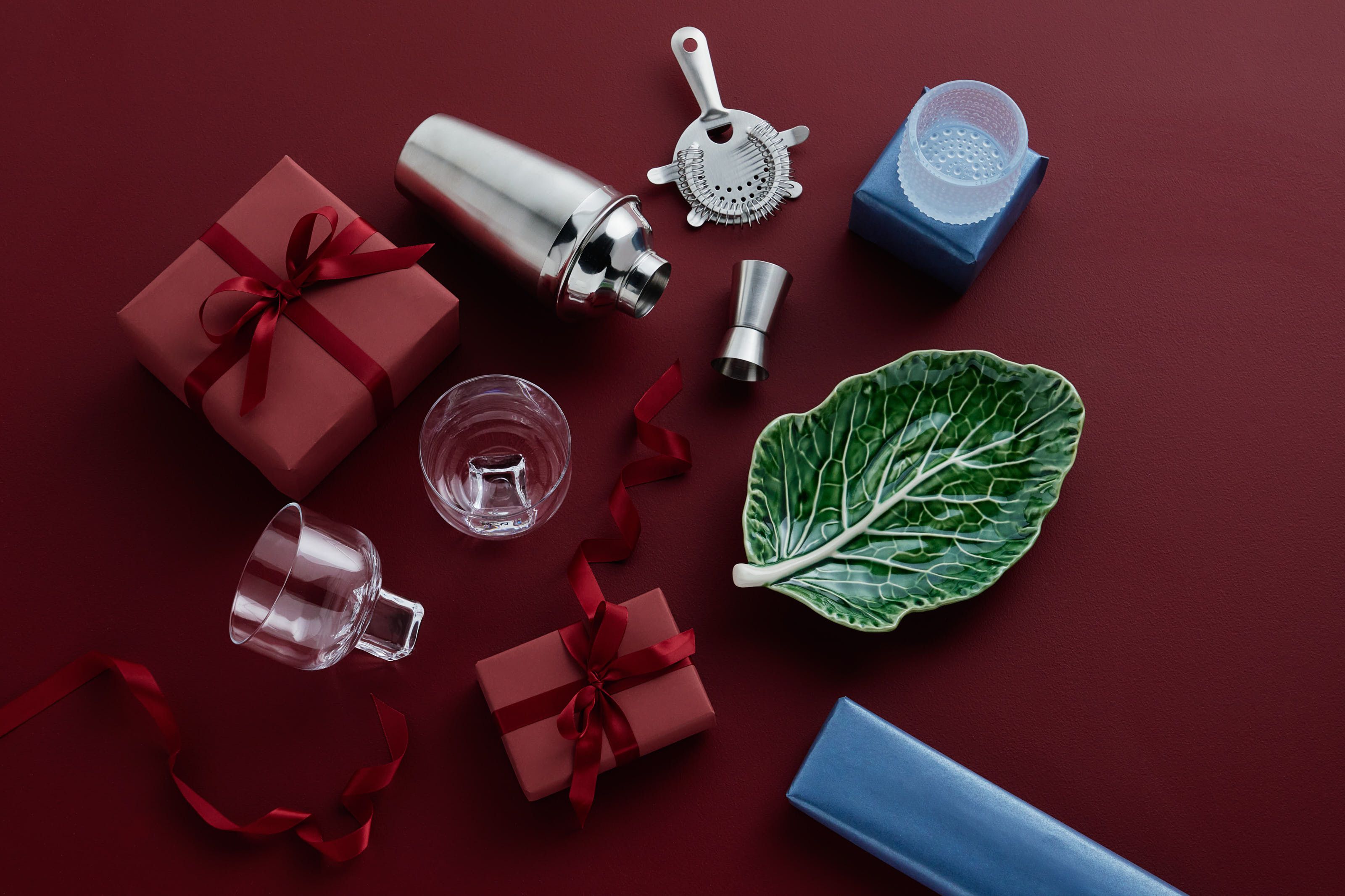 Cocktailshaker, barverktyg, glas, en bladtallrik och presentaskar i rött och blått på en mörkröd yta.