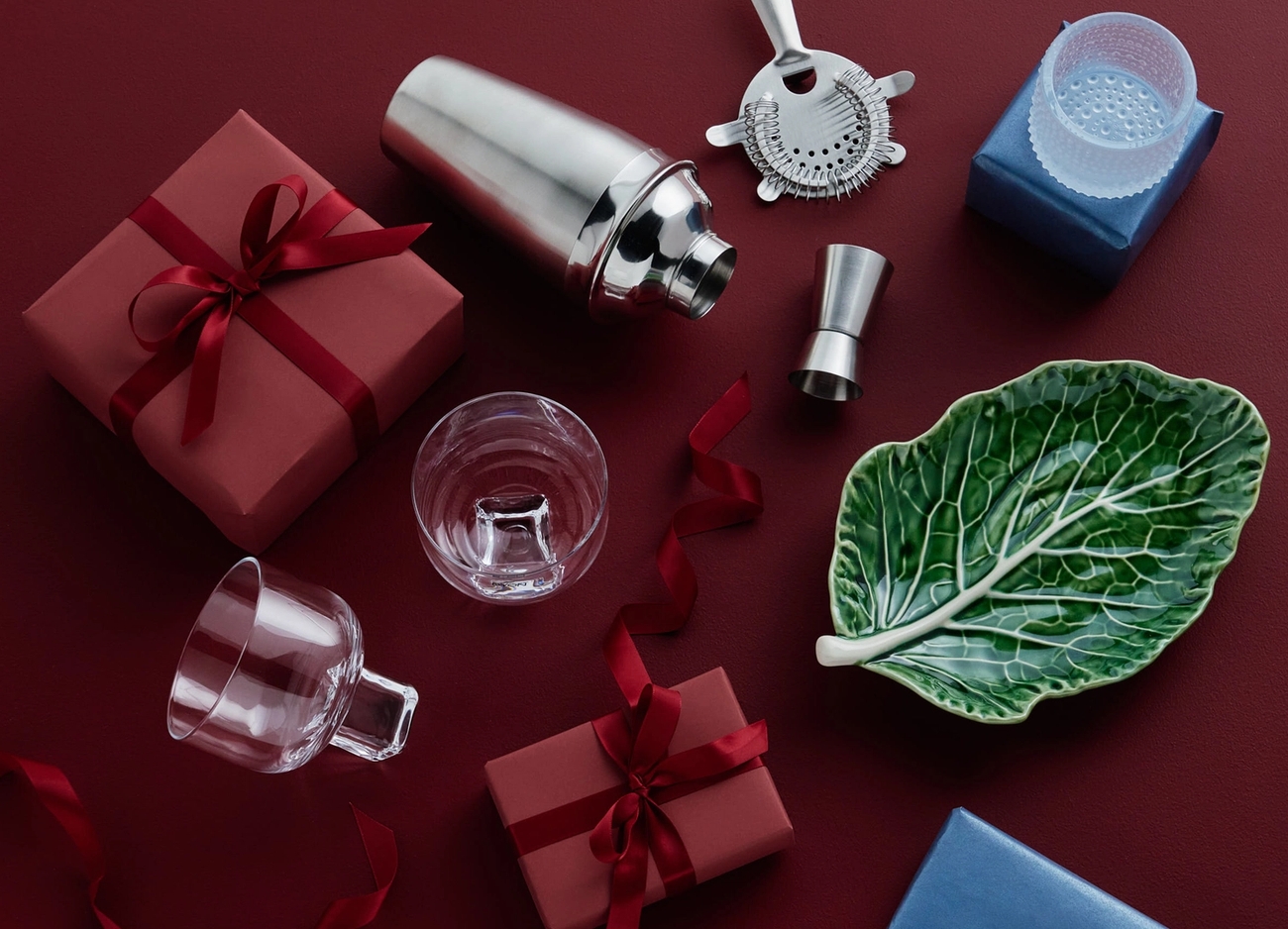 Cocktailshaker, barverktyg, glas, en bladtallrik och presentaskar i rött och blått på en mörkröd yta.