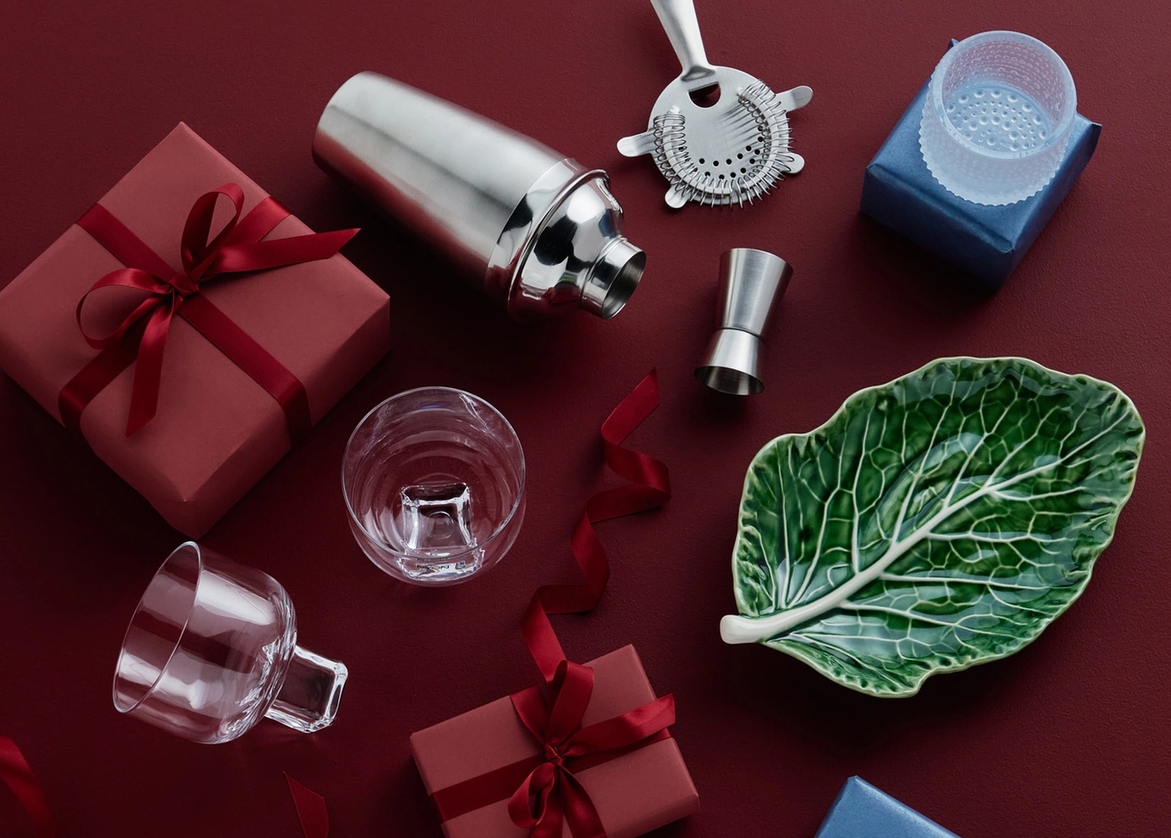 Cocktailshaker, barverktyg, glas, en bladtallrik och presentaskar i rött och blått på en mörkröd yta.