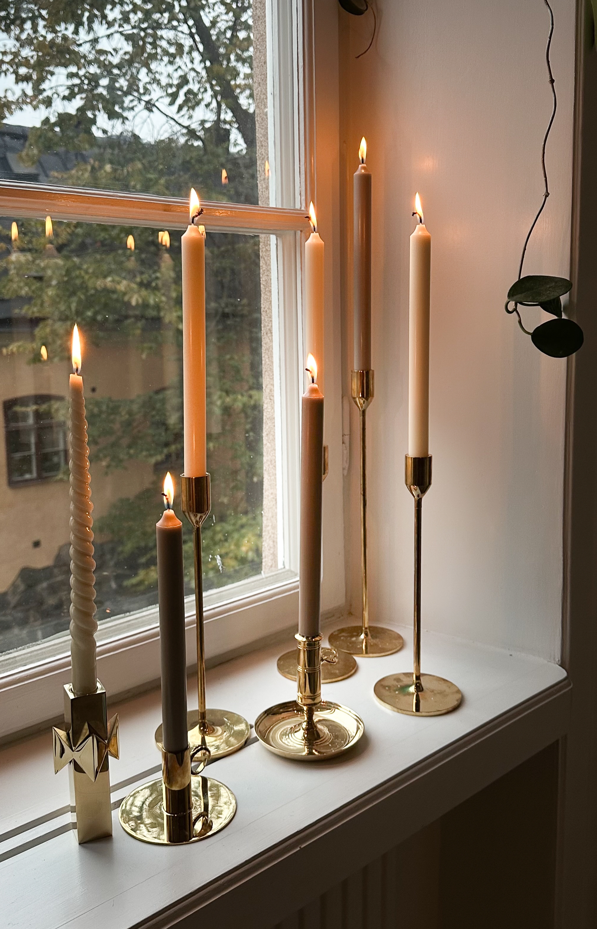 Seven lit candles in brass holders on a white windowsill, with a window showing trees outside.