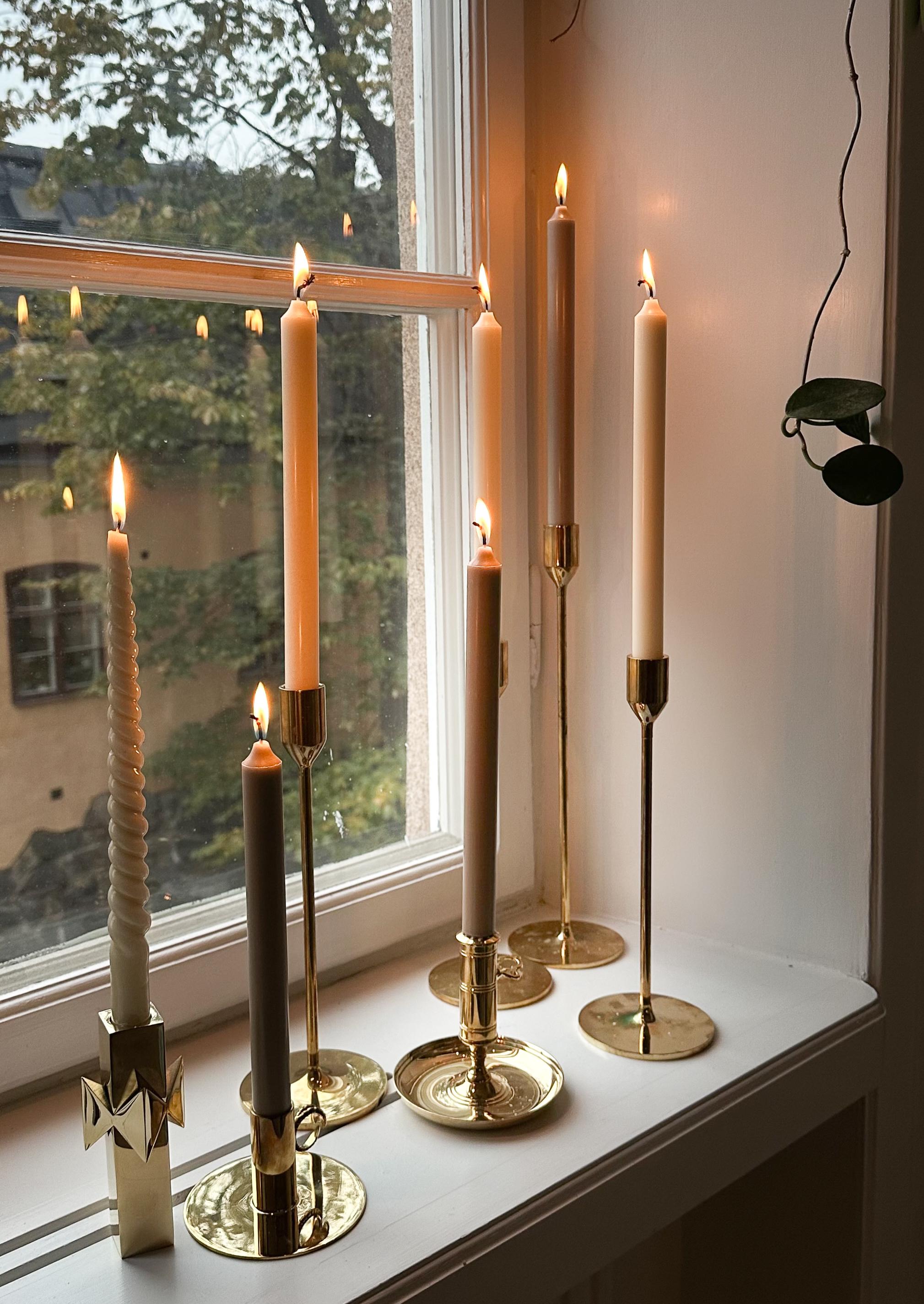 Seven lit candles in brass holders on a white windowsill, with a window showing trees outside.