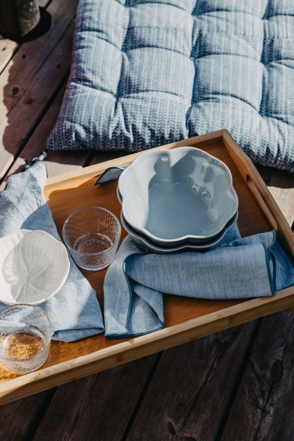 Et trebrett med lyseblå servise og linservietter står på en treterrasse ved siden av en mønstret blå pute