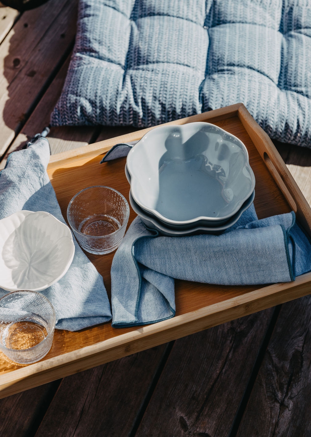 Et trebrett med lyseblå servise og linservietter står på en treterrasse ved siden av en mønstret blå pute