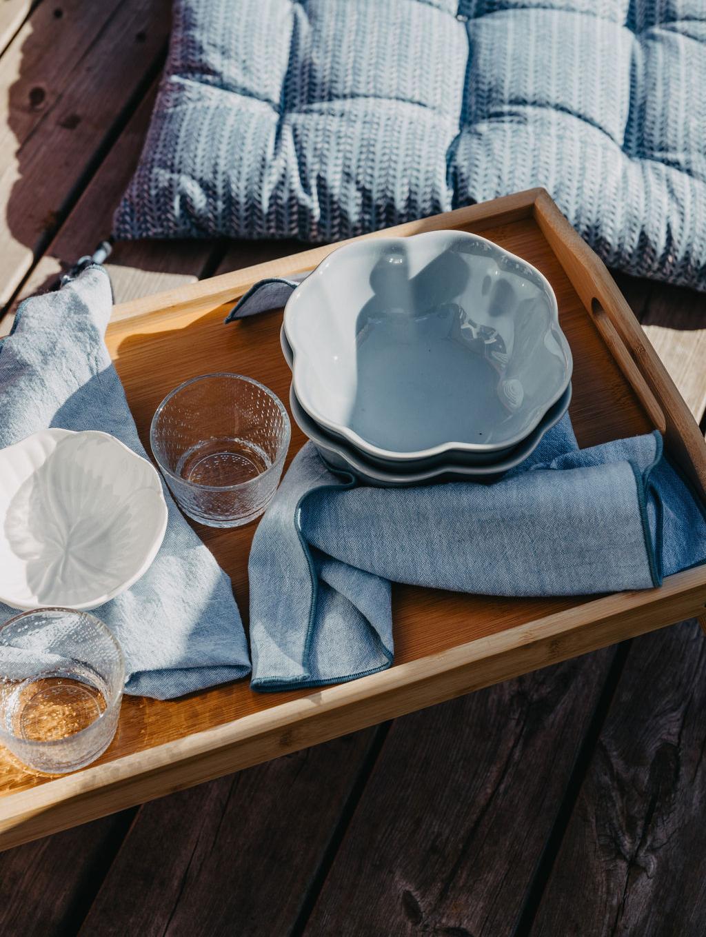 Et trebrett med lyseblå servise og linservietter står på en treterrasse ved siden av en mønstret blå pute