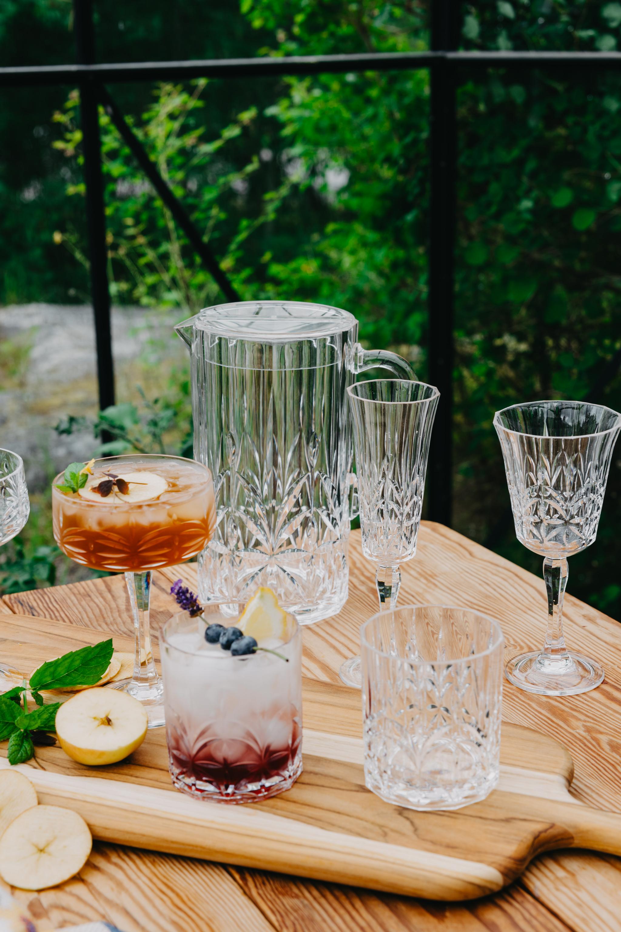 Eleganta kristallglas och karaff med somriga drinkar står på ett träbord i en grönskande trädgårdsmiljö.