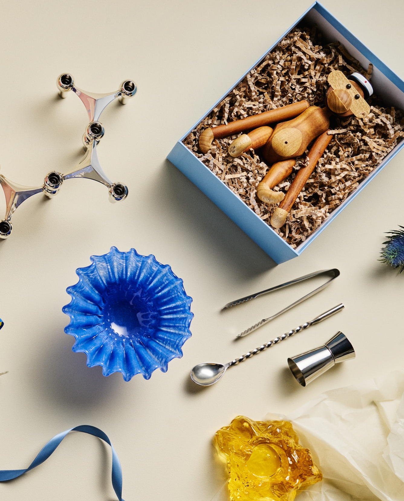 A wooden monkey toy in a blue gift box, a silver modular candle holder, a blue fluted glass bowl, and bar tools arranged on a light surface.
