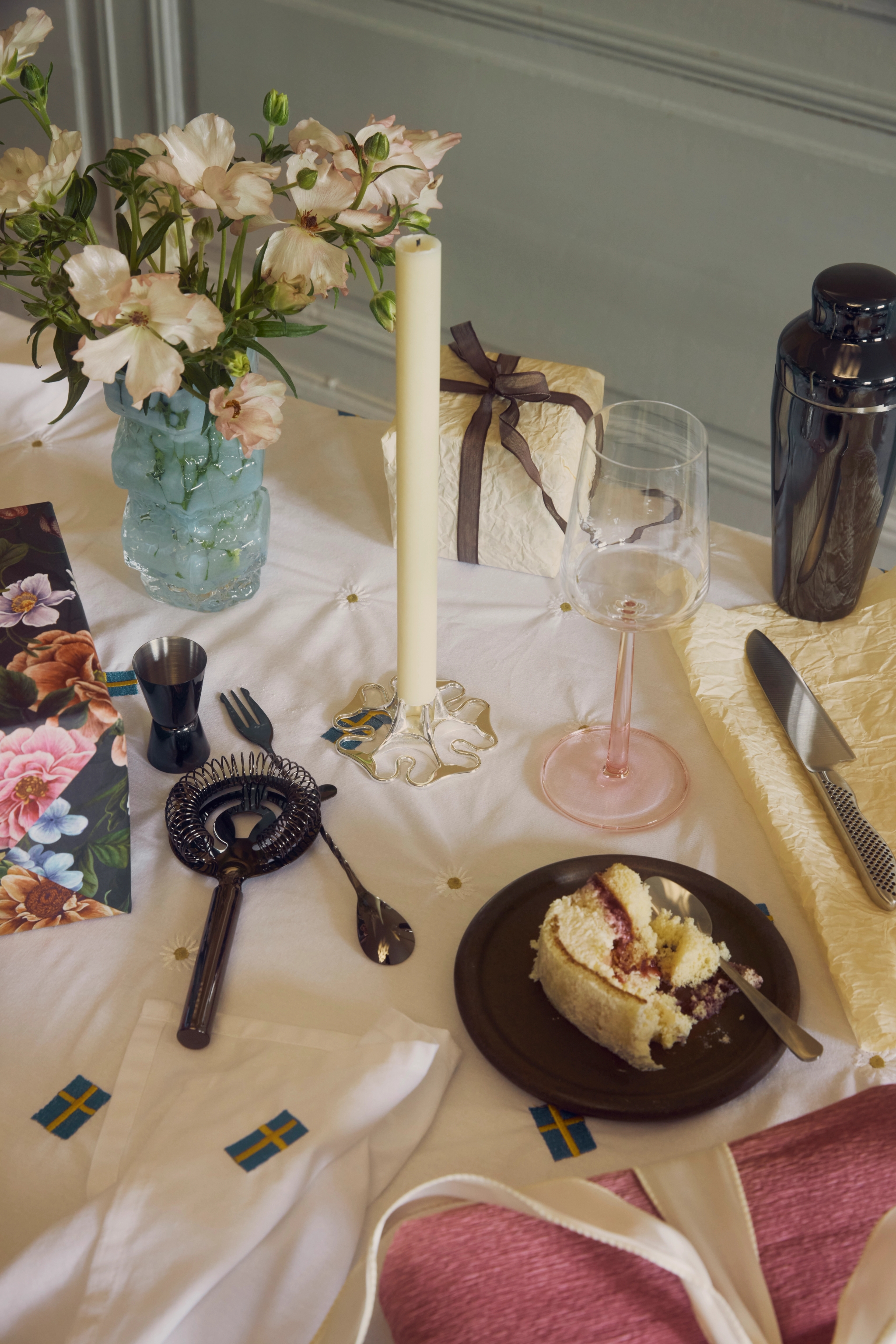 En dukning med rosa blommor, ett ljus, bartillbehör, en tårtbit, en present och detaljer med svenska flaggan på en vit duk.