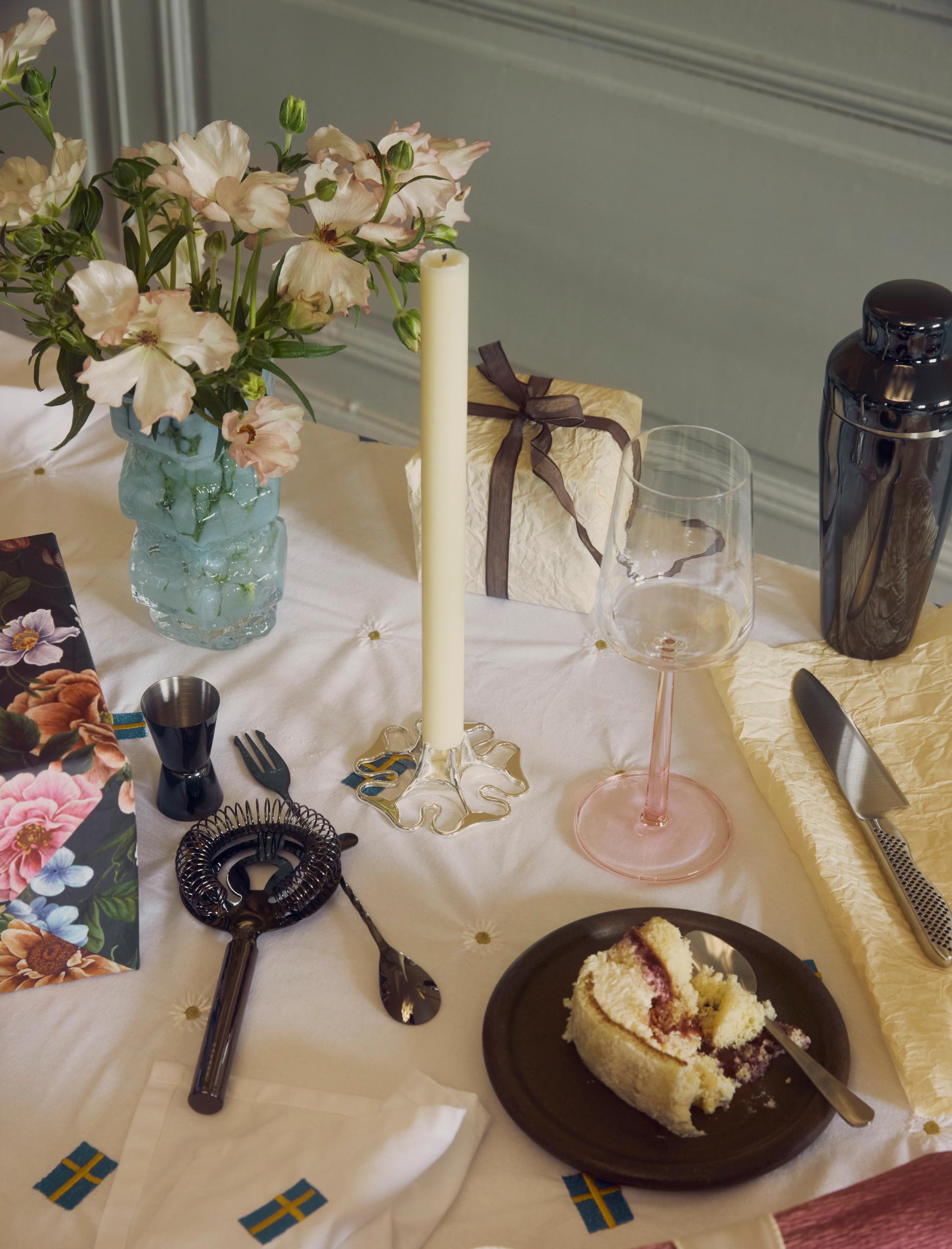En dukning med rosa blommor, ett ljus, bartillbehör, en tårtbit, en present och detaljer med svenska flaggan på en vit duk.