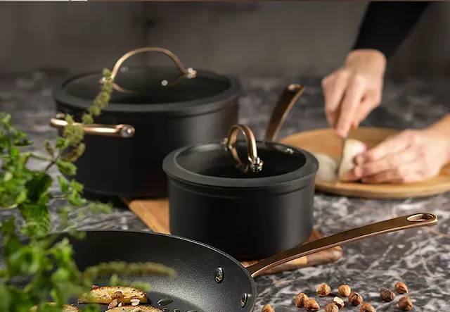 Tyylikkäät mustat kattilat ja paistinpannu keittiön marmoritasolla, jossa paistetaan perunoita ja pilkotaan pähkinöitä.