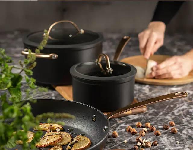 Tyylikkäät mustat kattilat ja paistinpannu keittiön marmoritasolla, jossa valmistetaan ruokaa ja pilkotaan aineksia.
