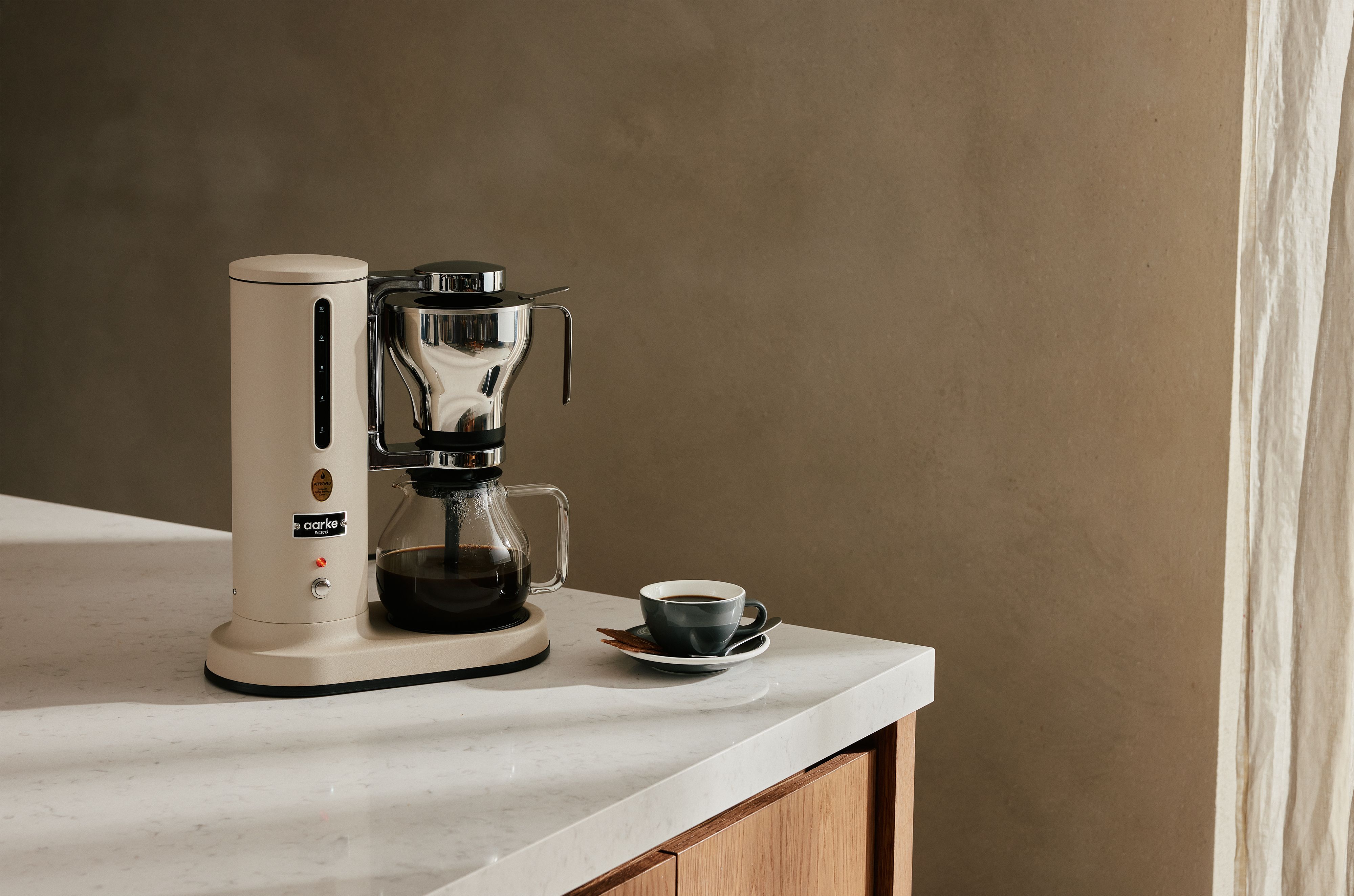 Stilren kaffetrakter i beige og stål står på en lys kjøkkenbenk ved siden av en kopp nybrygget kaffe.