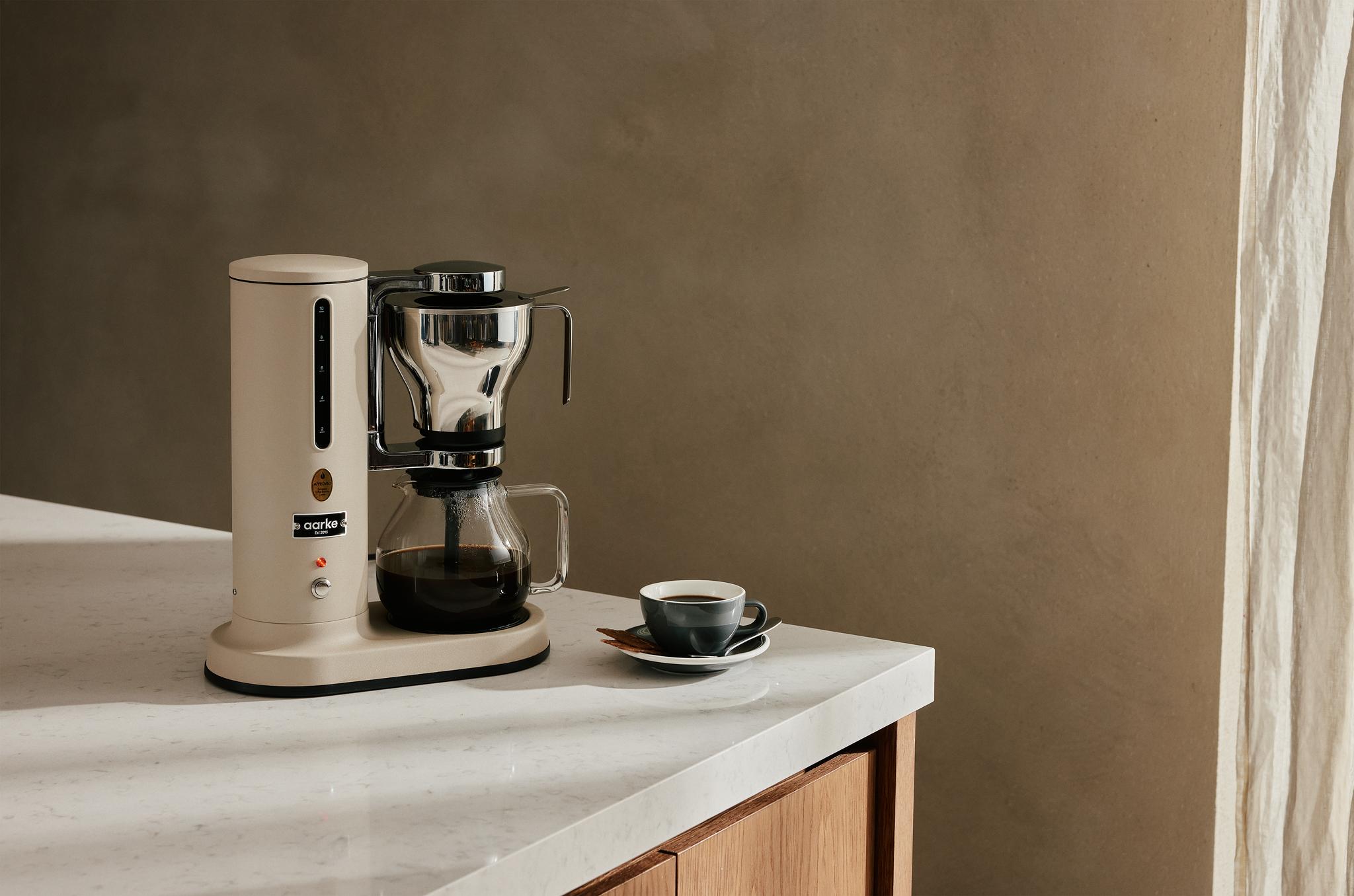Elegant kaffebryggare i rostfritt stål och beige står på en marmorbänk bredvid en kopp nybryggt kaffe i ett stilrent kök.
