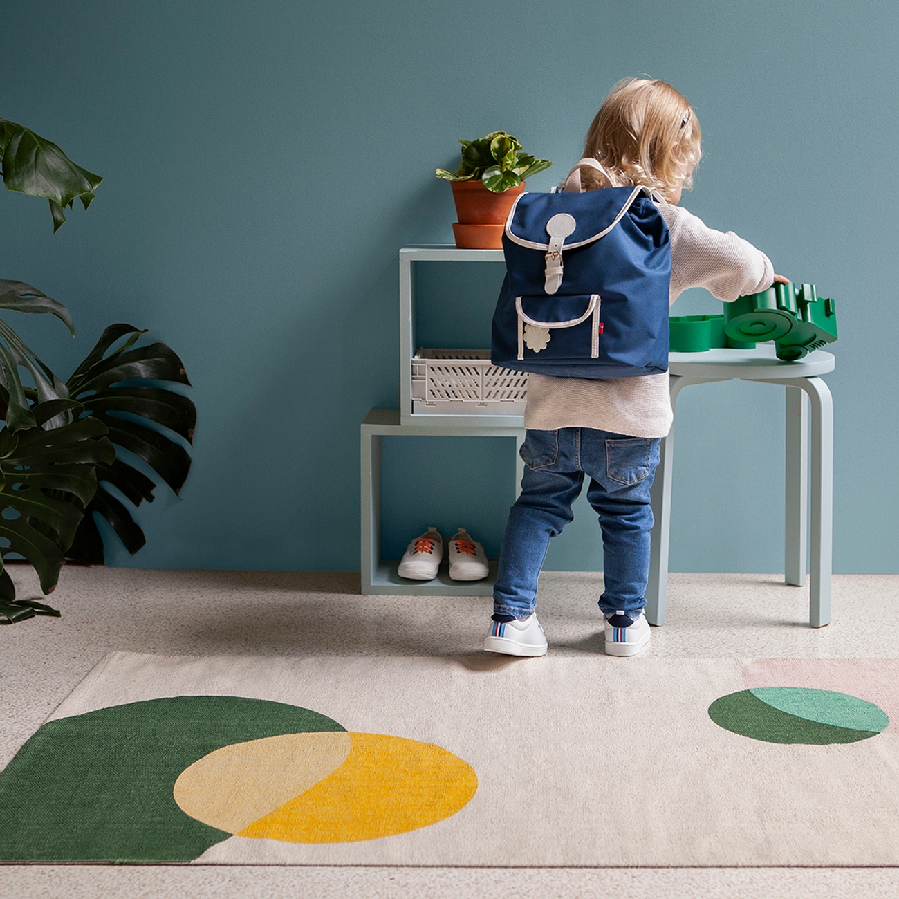 A toddler wearing a blue backpack plays at a light blue table in a room with a blue wall and a geometric rug.