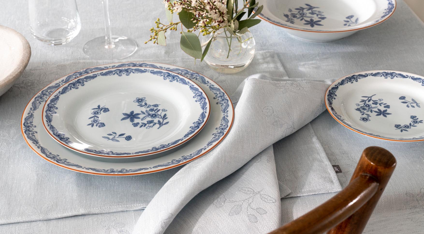 Ett matbord dukat med vita tallrikar med blå blomstermönster, ljusblått linnetyg och en liten vas med blommor.