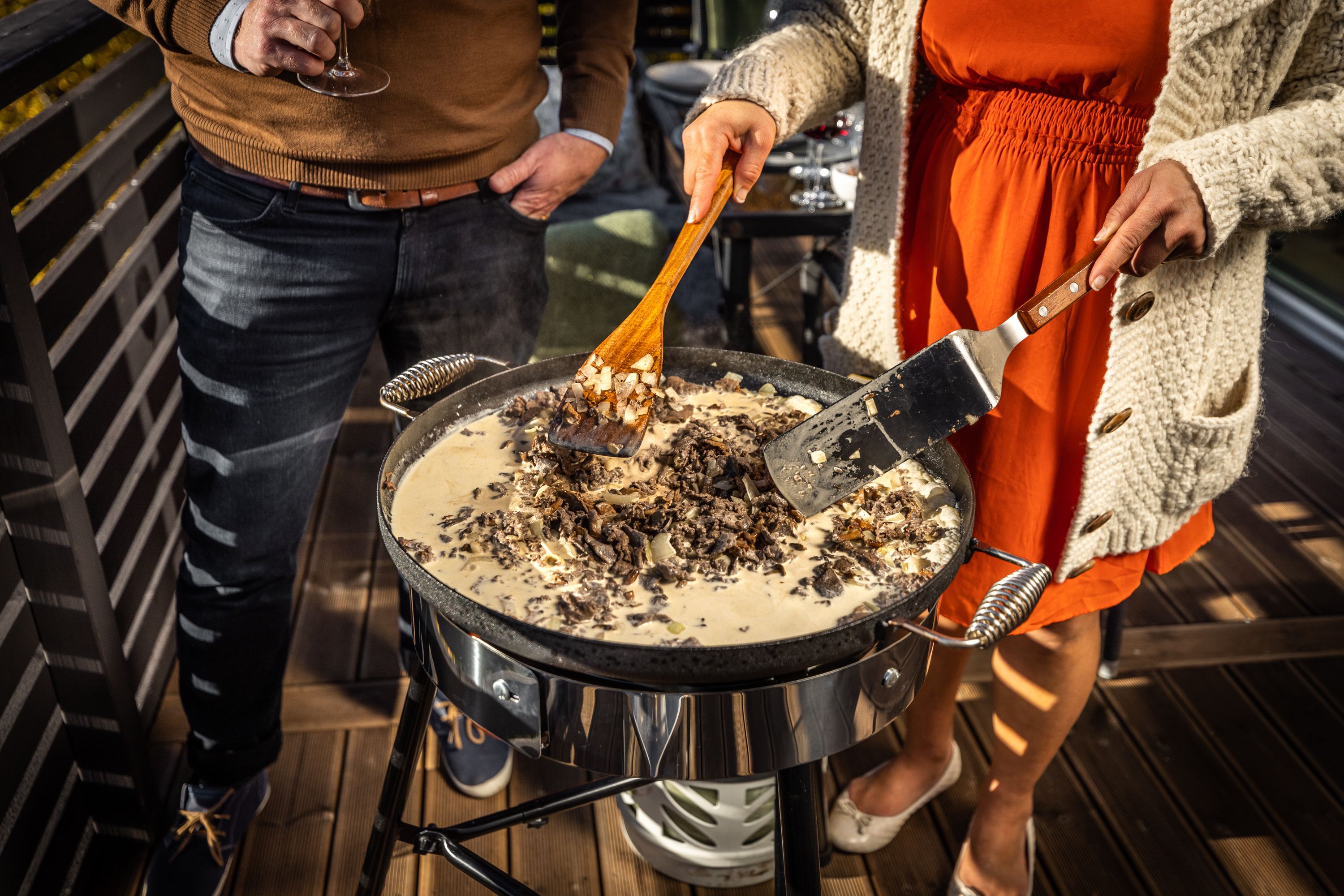 En kvinna rör om i en krämig rätt som lagas i en stor panna på en utomhusgrill, medan en man håller ett vinglas i närheten