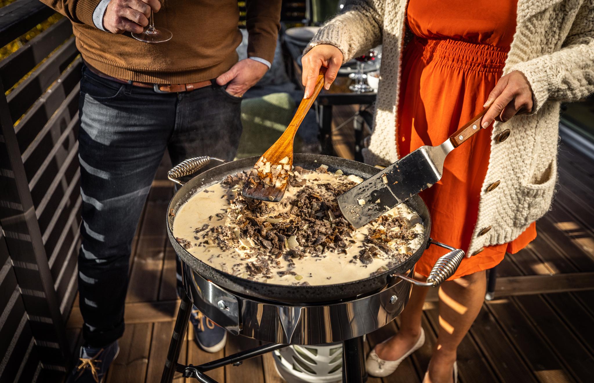 En kvinna rör om i en krämig rätt som lagas i en stor panna på en utomhusgrill, medan en man håller ett vinglas i närheten