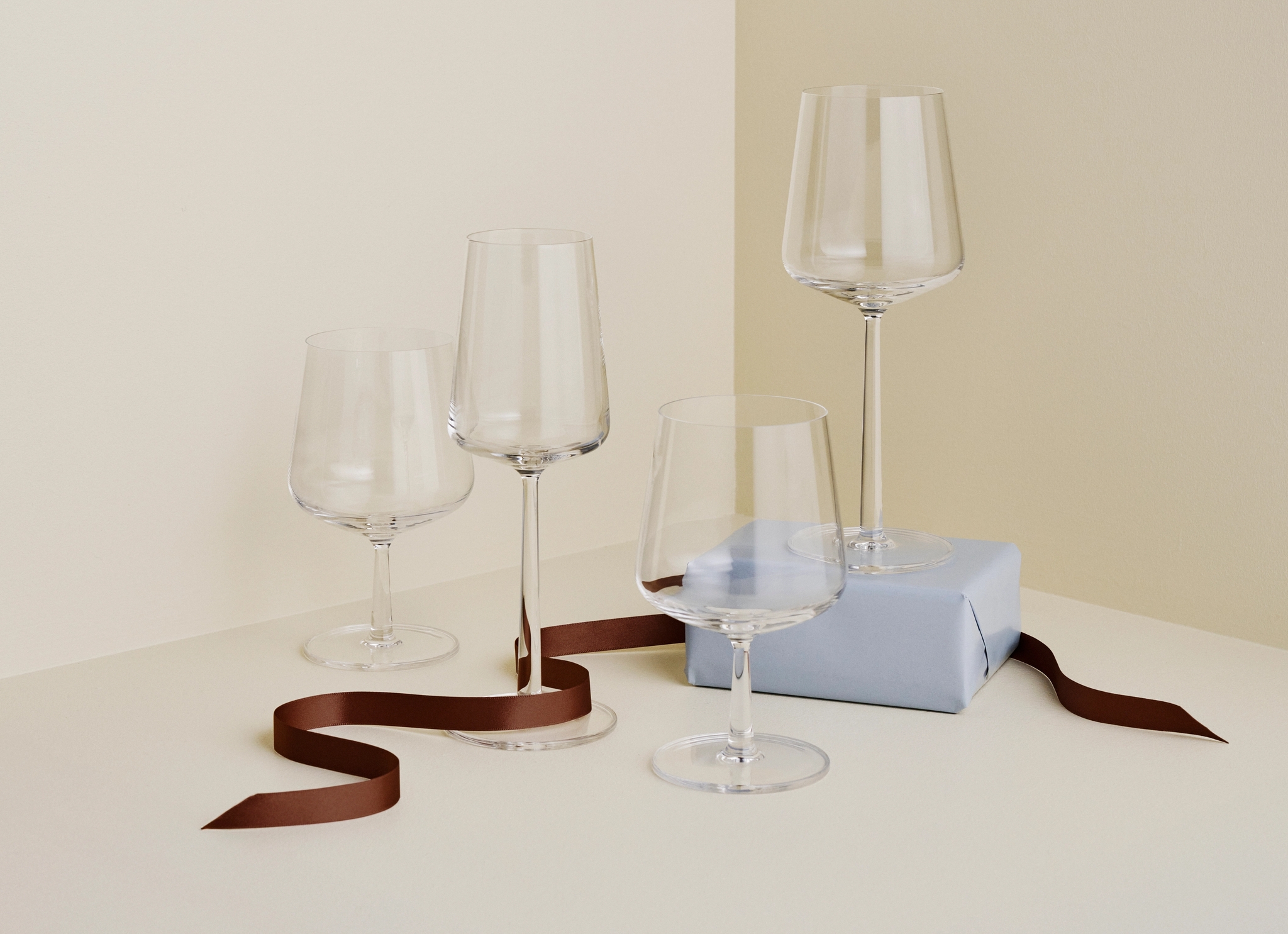 Fyra eleganta vinglas står arrangerade på ett bord med ett ljusblått paket och ett brunt sidenband mot en beige minimalistisk bakgrund.