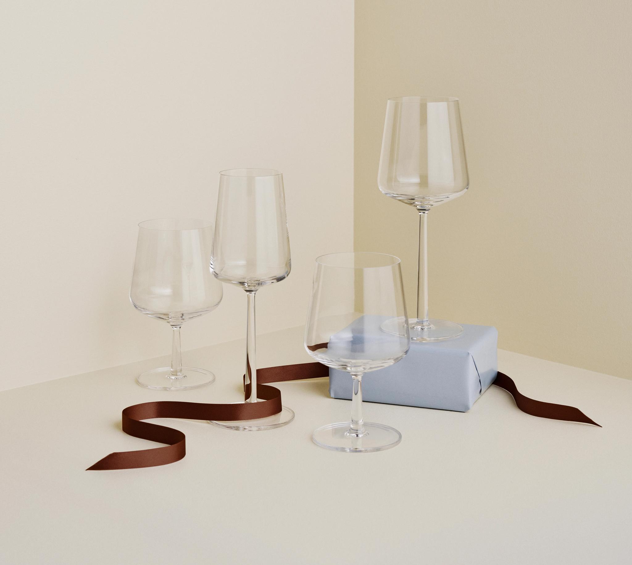 Fire elegante vinglass står på et lyst bord ved siden av en lyseblå gaveeske og et brunt silkebånd i et minimalistisk interiør.