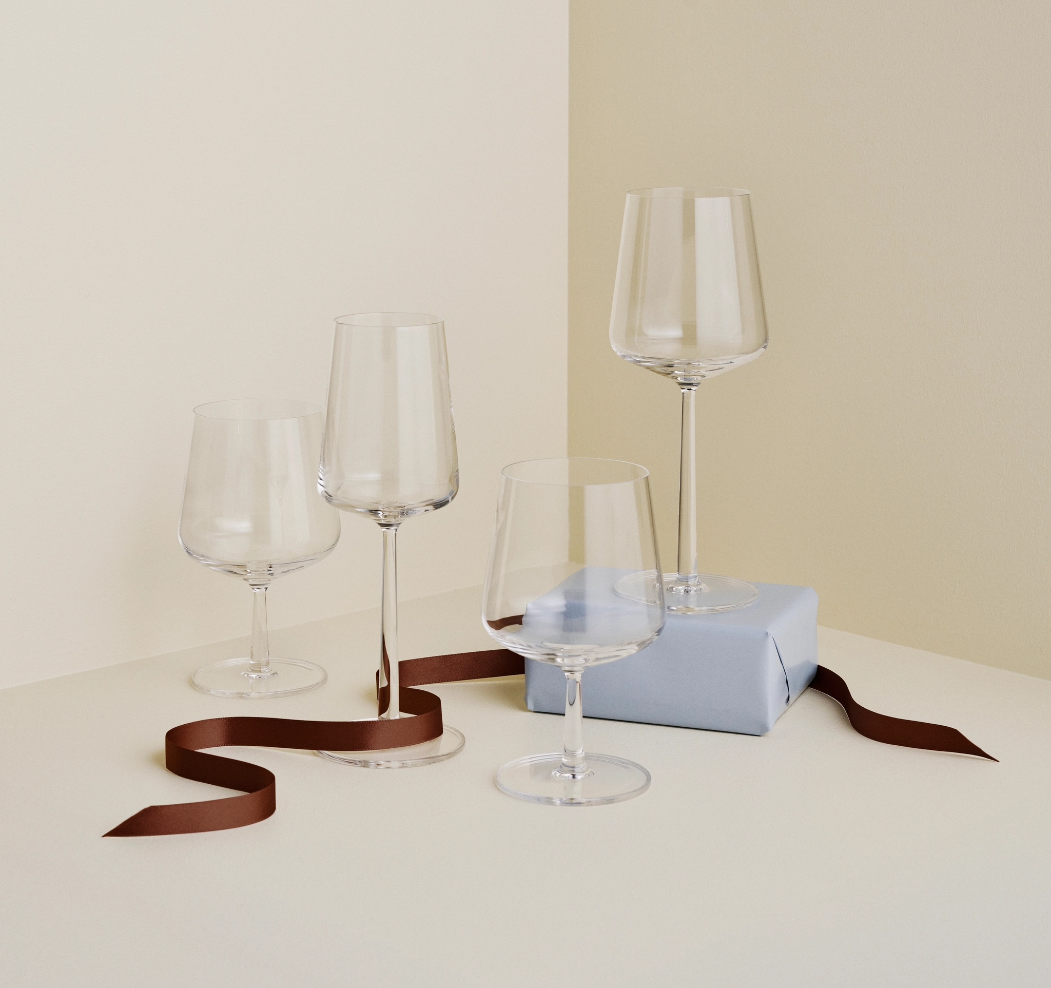 Fire elegante vinglass står på et lyst bord ved siden av en lyseblå gaveeske og et brunt silkebånd i et minimalistisk interiør.