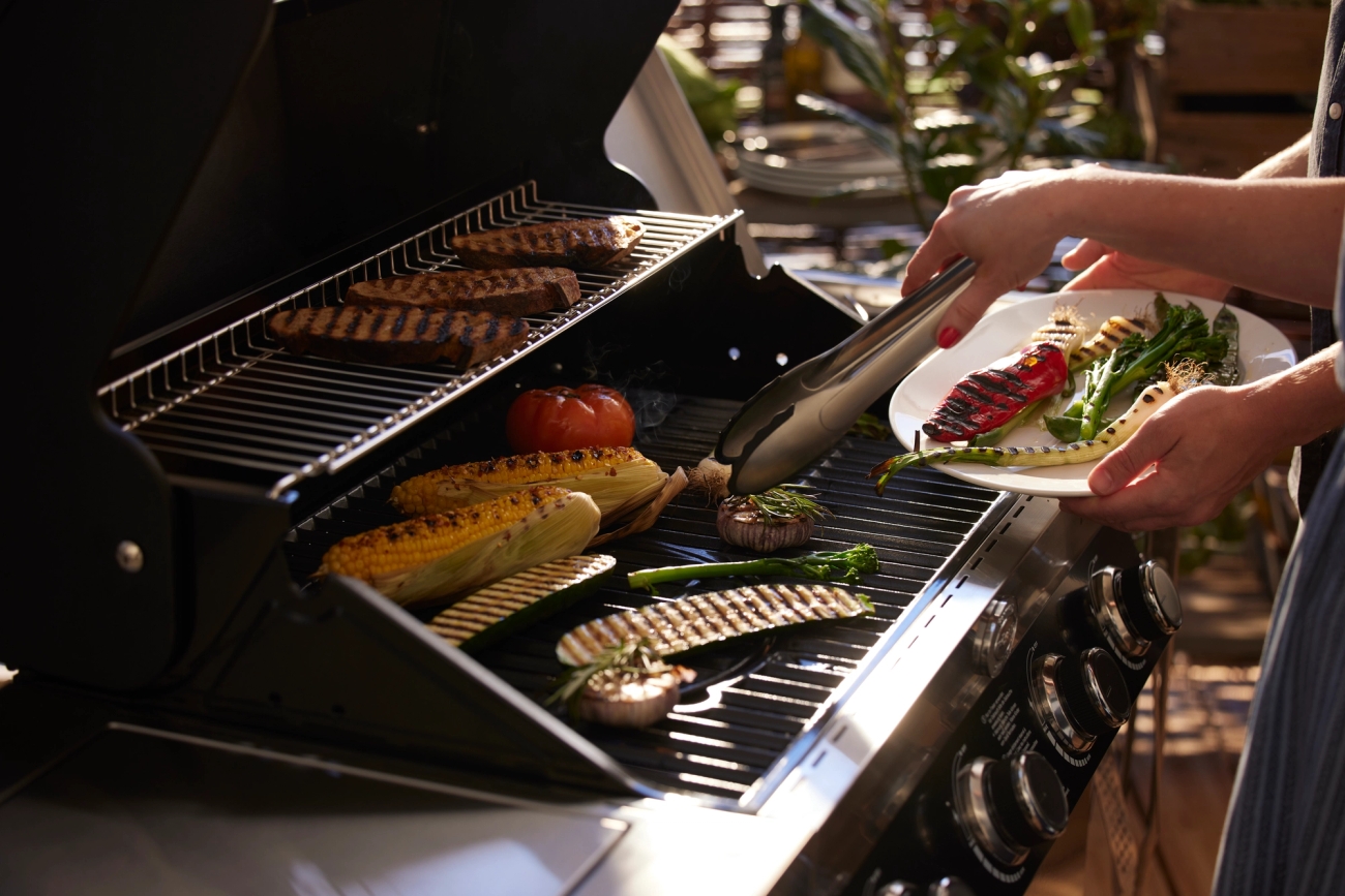 En person lägger upp grillade grönsaker på en tallrik medan andra grönsaker och kött tillagas på en gasolgrill