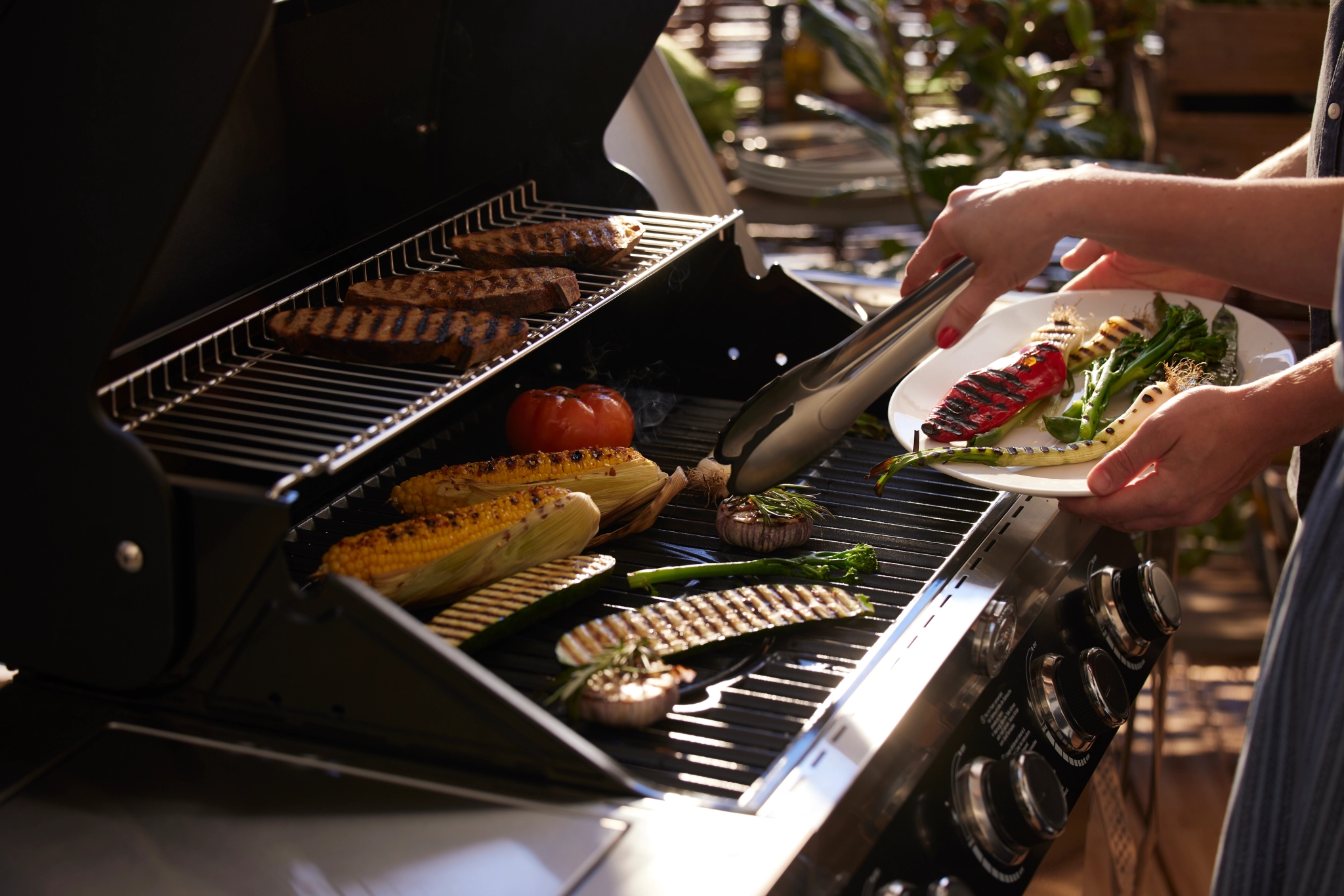 En person använder en tång för att lägga grillade grönsaker på en tallrik från en grill och tillaga olika maträtter.