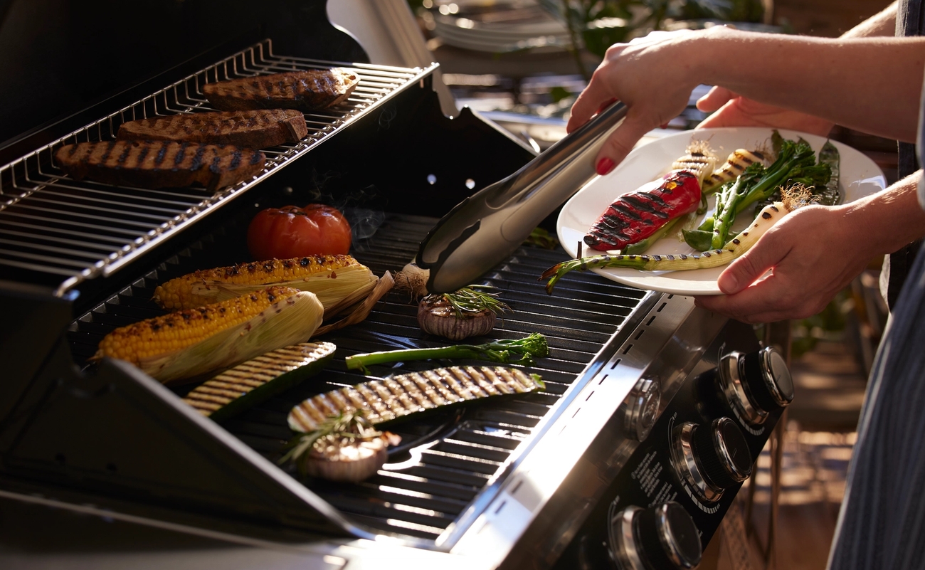 En person lägger grillade grönsaker på en tallrik från en grill där kött, majs och andra grönsaker tillagas