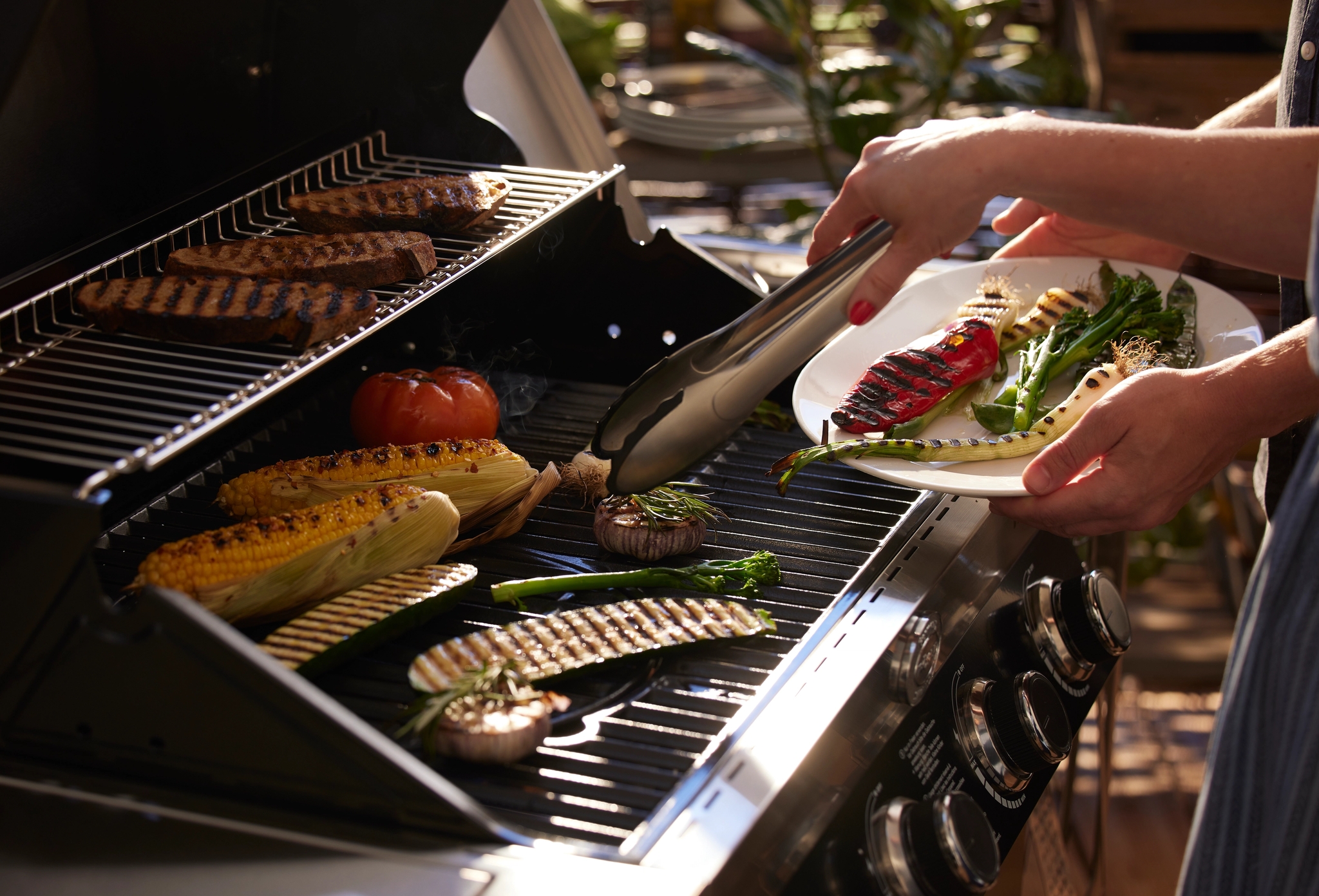 En person legger grillede grønnsaker på en tallerken fra en grill som tilbereder kjøtt, mais og andre grønnsaker