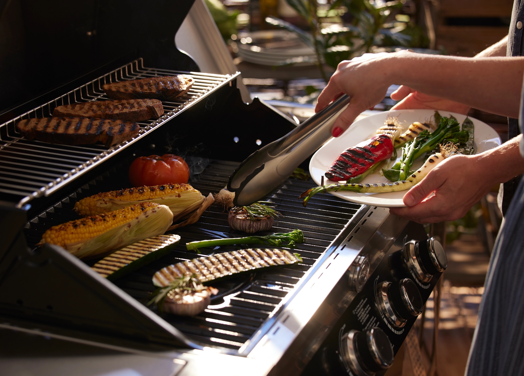 Henkilö siirtää grillattuja vihanneksia lautaselle grillistä, jossa kypsyy lihaa, maissia ja muita vihanneksia