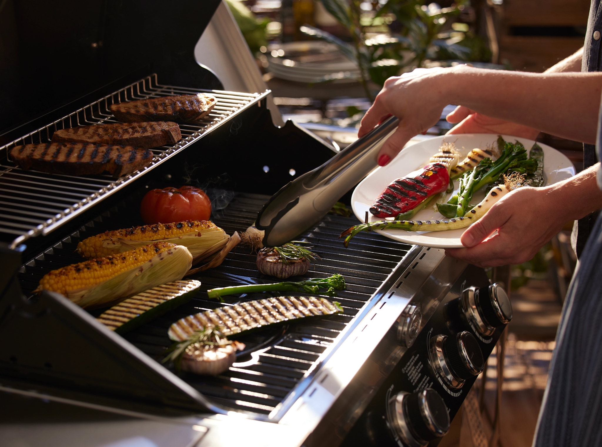 Henkilö siirtää grillattuja vihanneksia lautaselle grillistä, jossa kypsyy lihaa, maissia ja muita vihanneksia